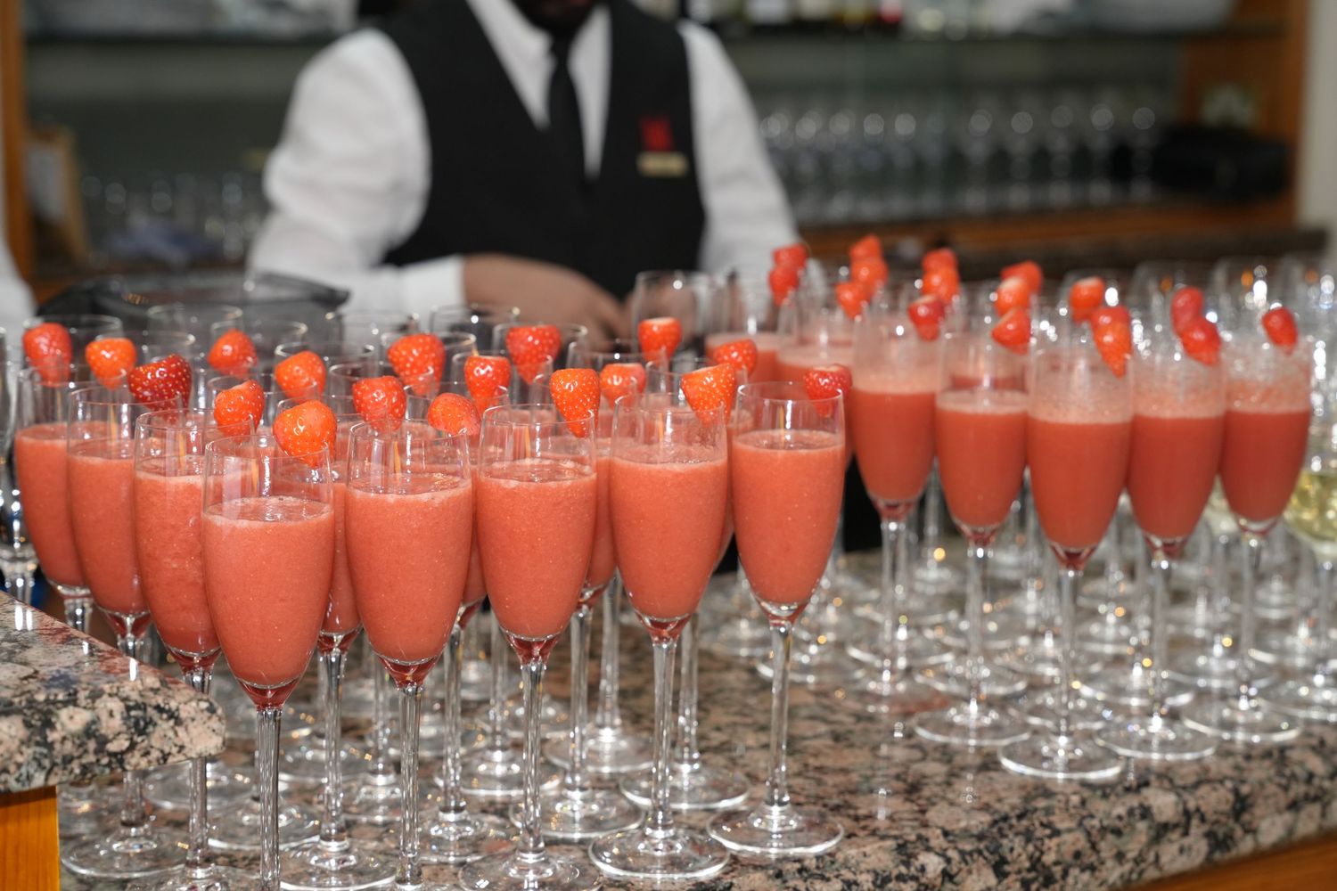 Rows of elegant champagne flutes filled with pink strawberry cocktails garnished with fresh strawberries served by a bartender.