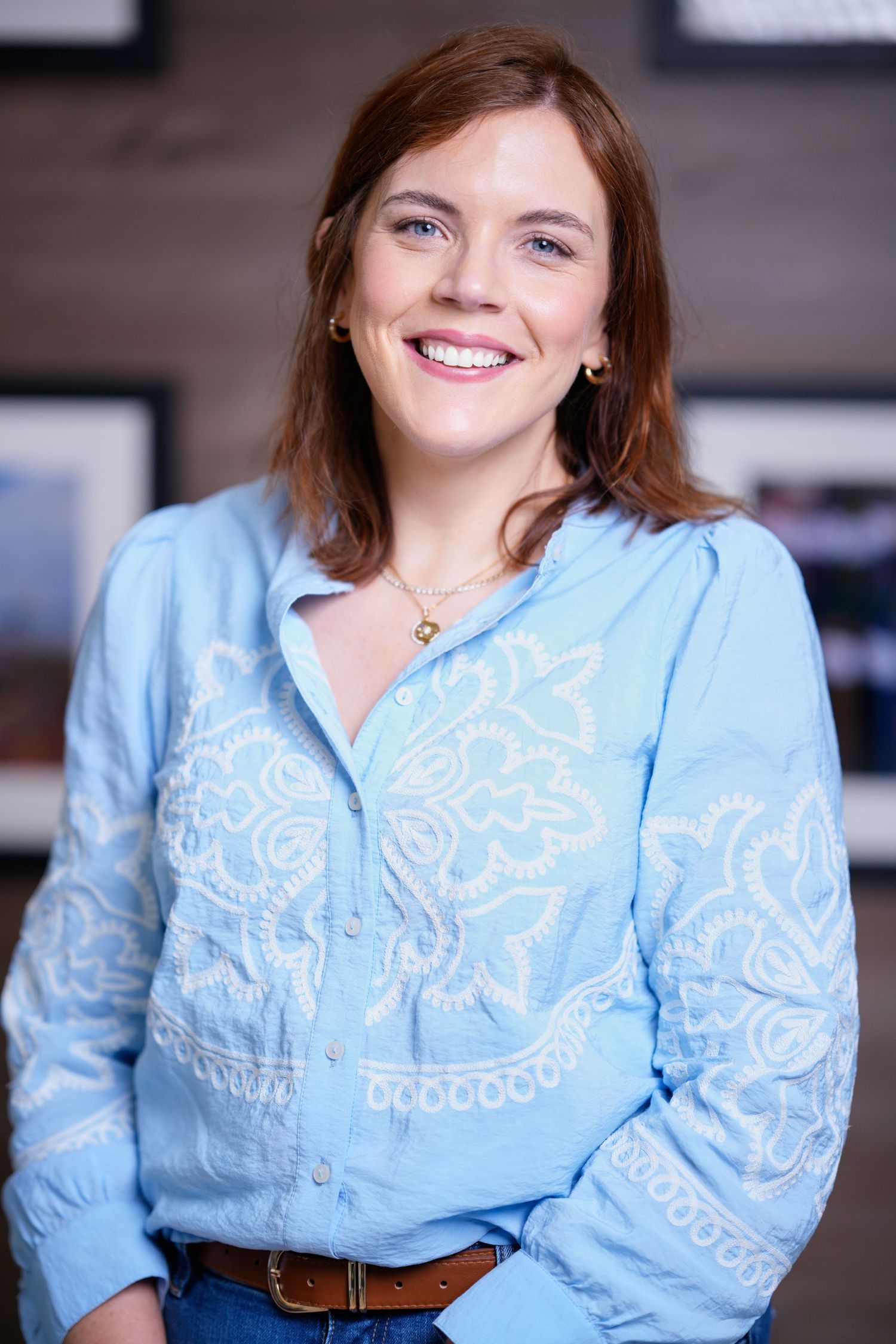 Smiling woman in light blue embroidered blouse and jeans, wearing a gold necklace, posing indoors.