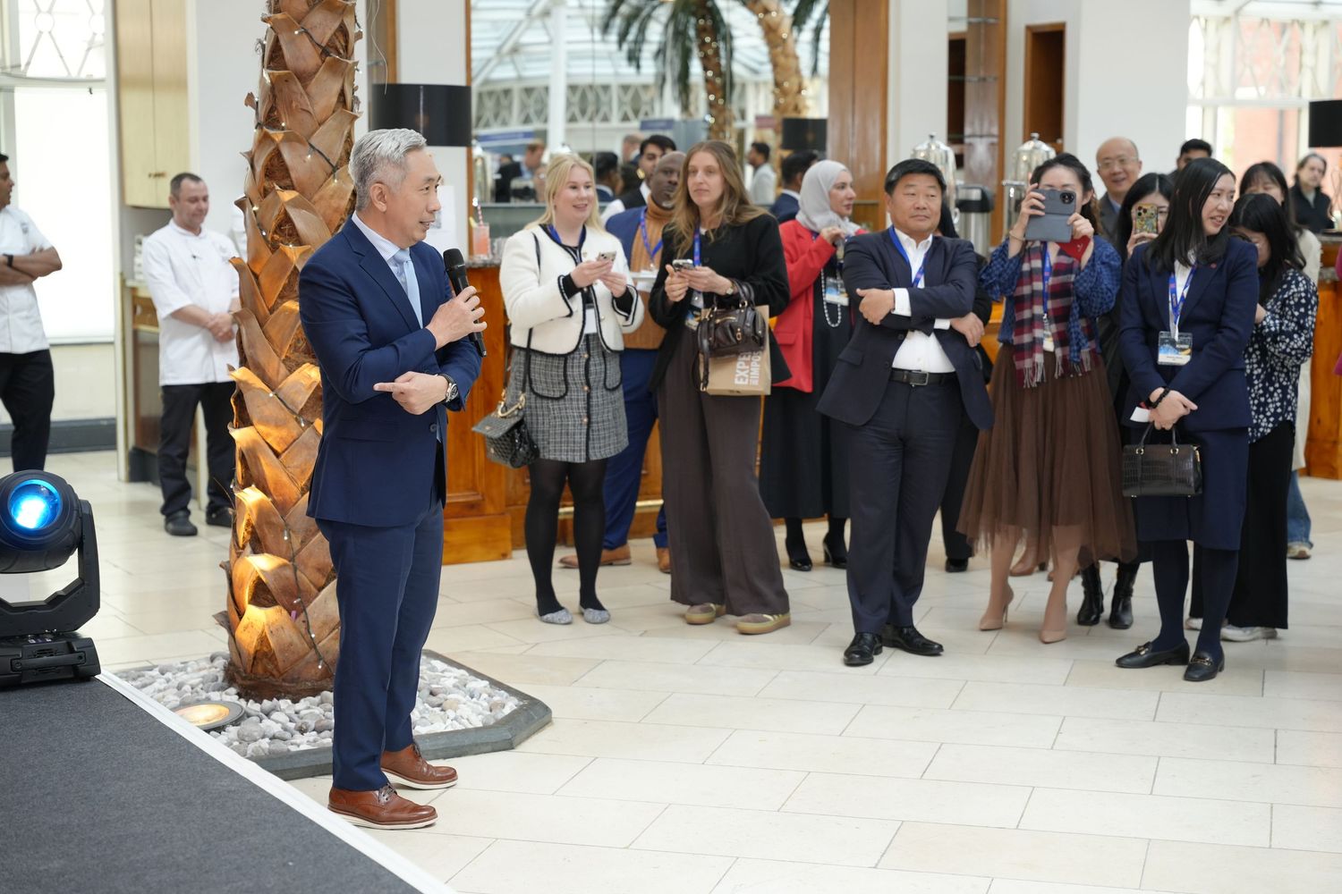A speaker addresses a diverse group of attendees at an indoor corporate event in a modern building lobby.