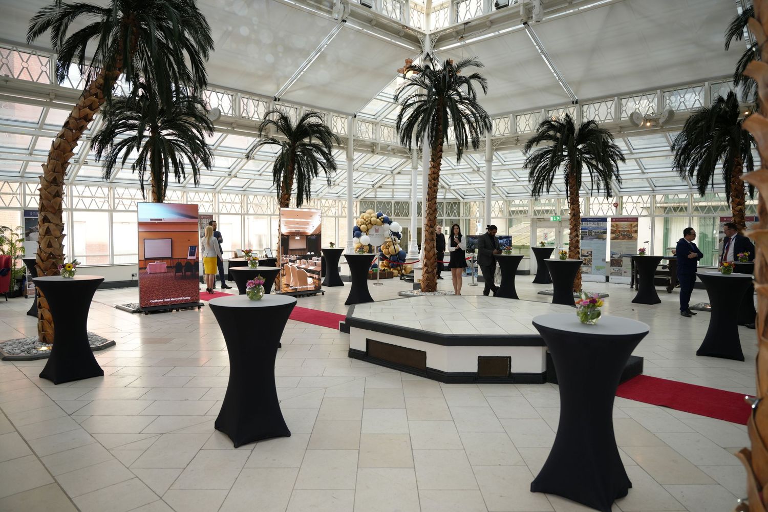 Elegant event hall with artificial palm trees, black cocktail tables, and guests mingling under a glass ceiling.