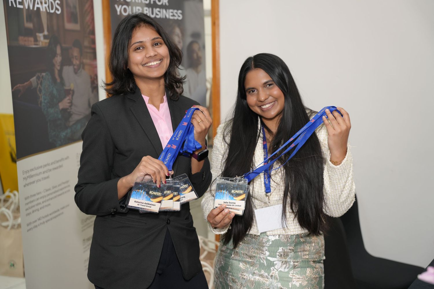 Two women smiling at a business event, holding blue lanyards and promotional materials at a corporate conference.