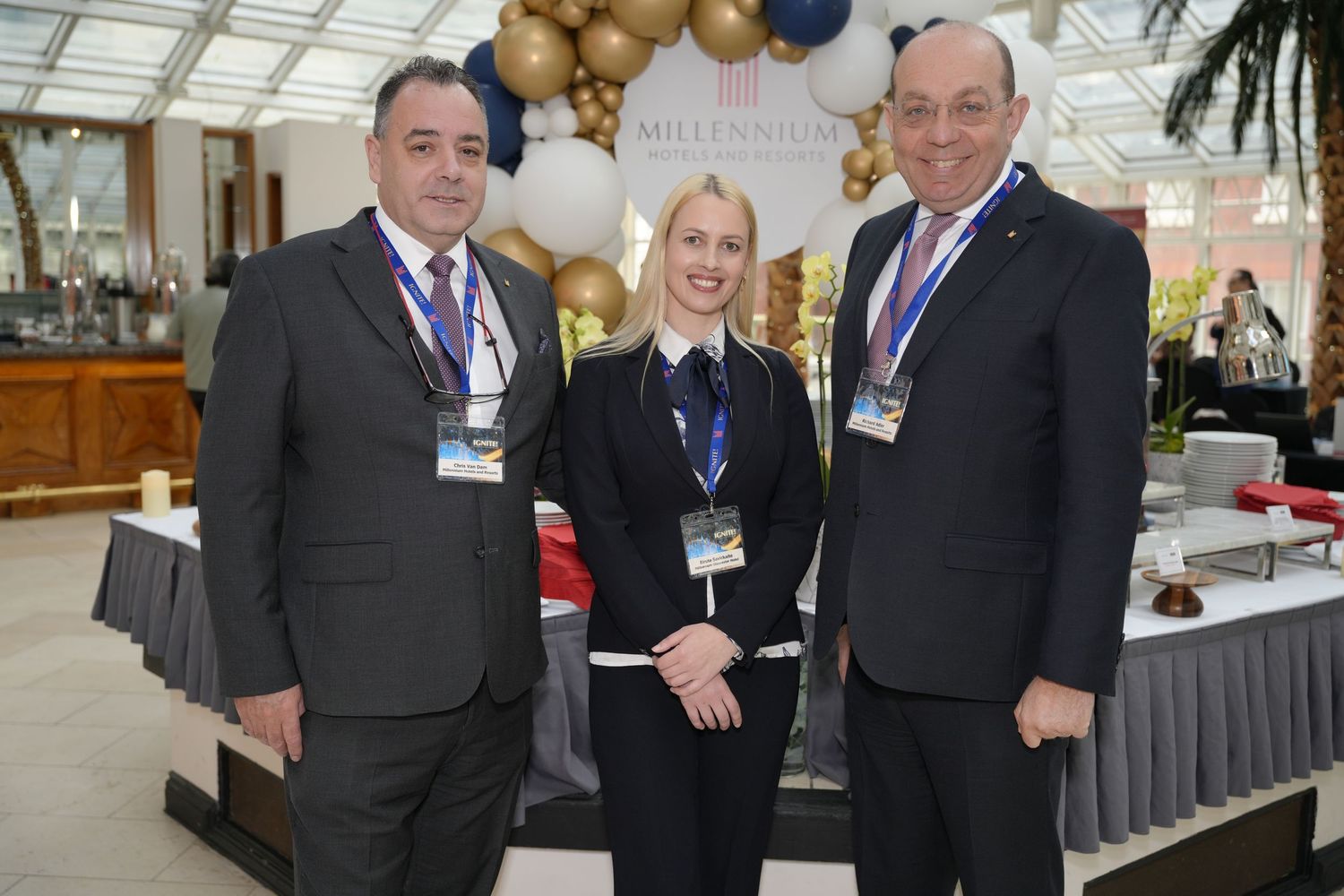 Three professionals in formal attire smile at a Millennium Hotels event with balloons and decor in the background.
