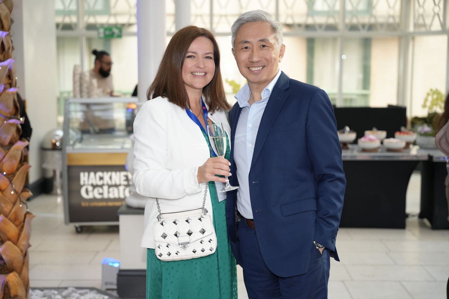 A smiling couple poses together at a social event, holding champagne glasses near a Hackney Gelato stand.