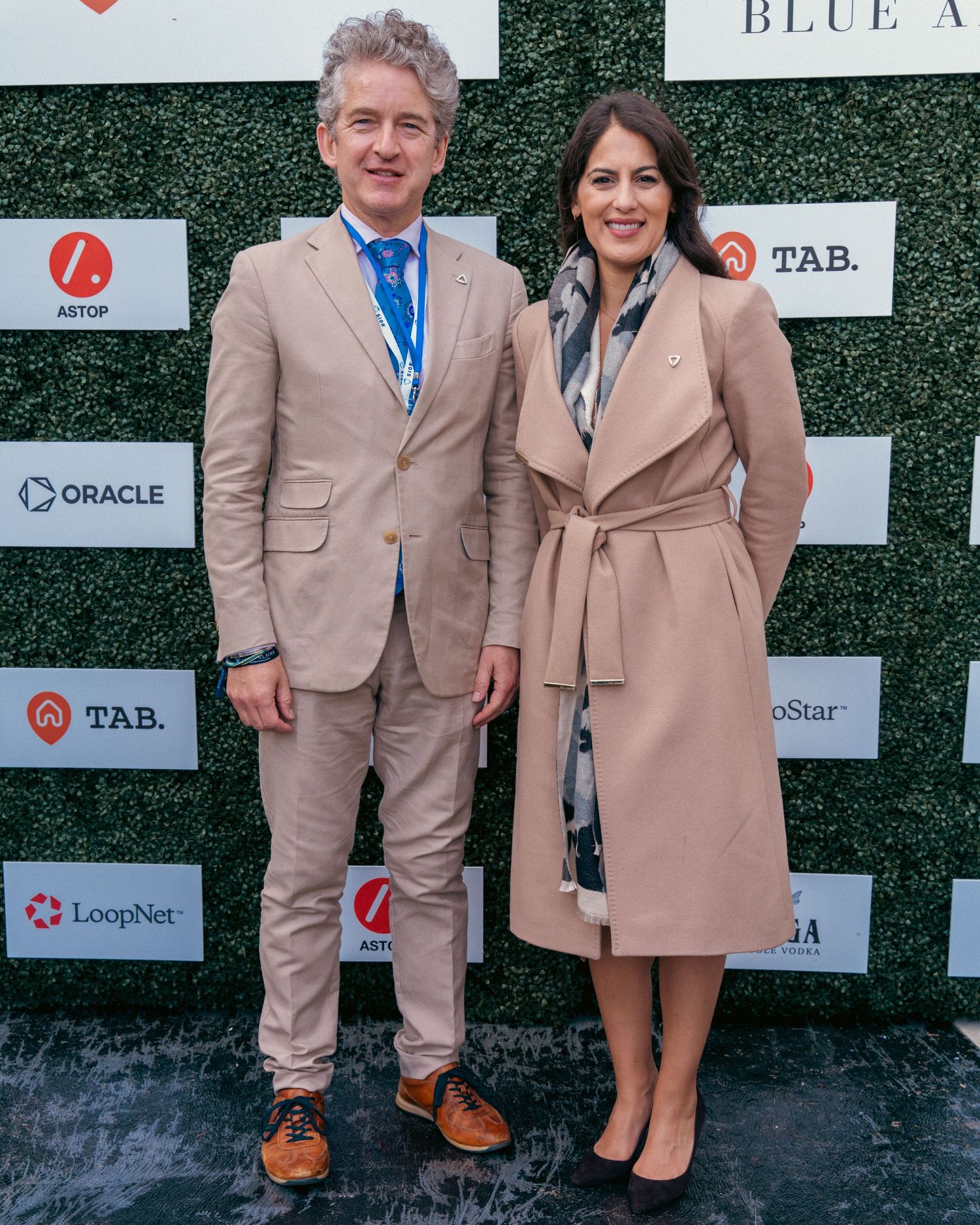 Two people in matching beige suits posing against branded step-and-repeat wall.
