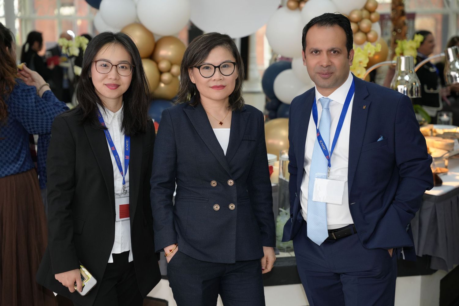 Three professionals in business attire wearing lanyards pose together at a corporate event with decorative balloons.