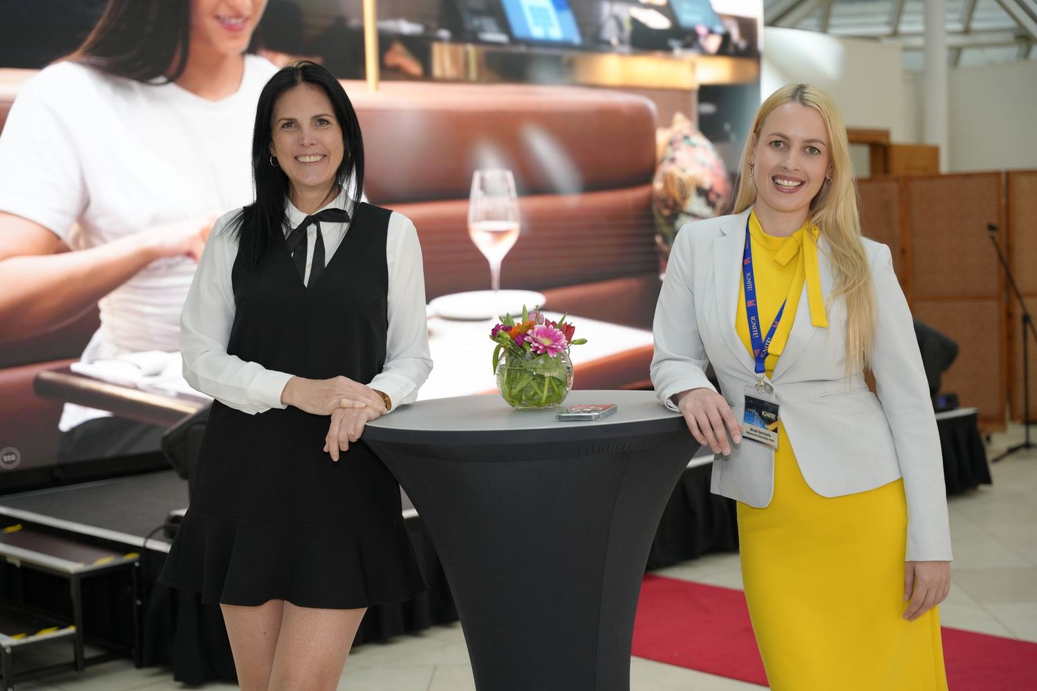 Two professional women smiling at a cocktail table at a corporate trade show event, one in black and one in yellow.