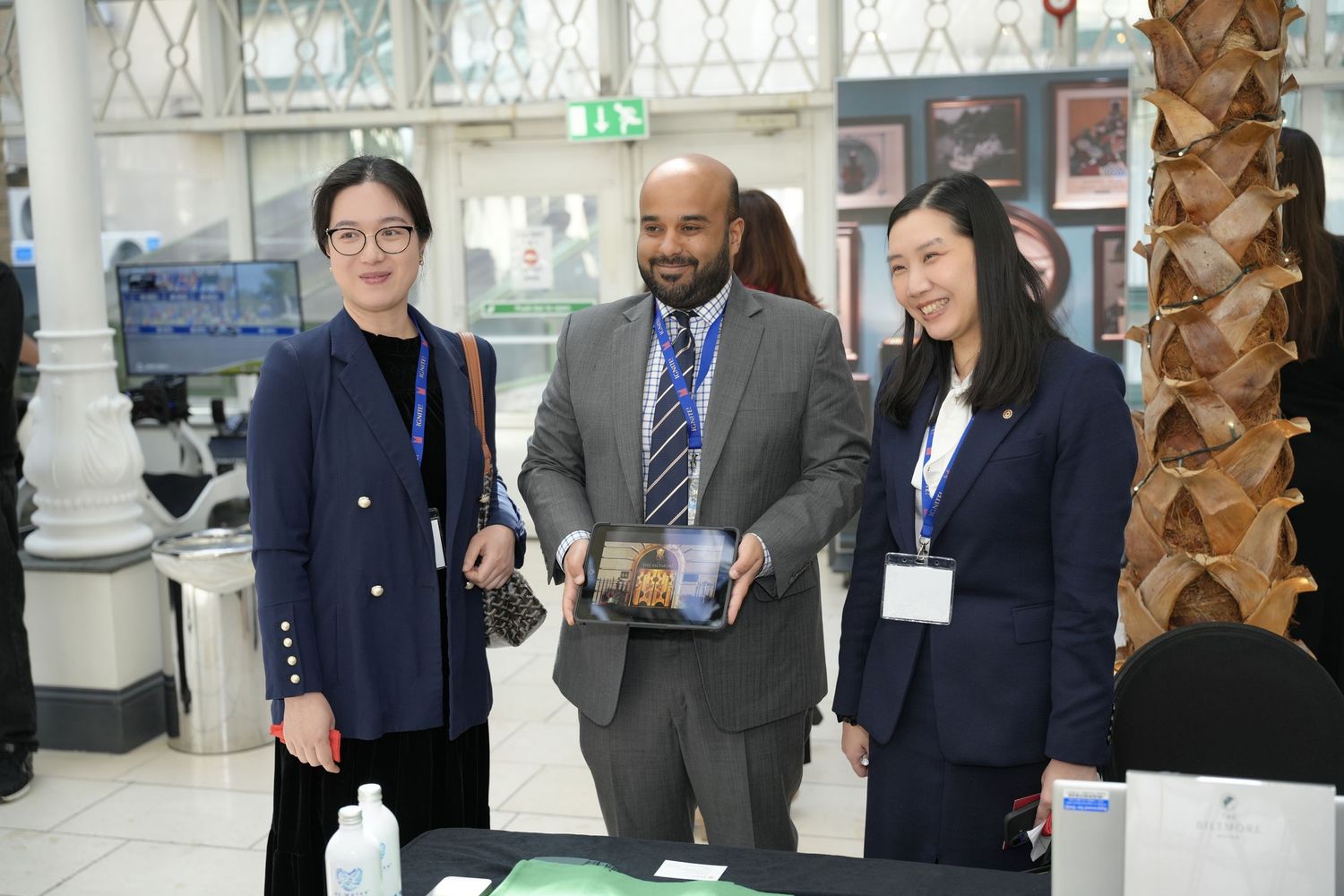 Three professionals stand together at a conference table, with one man holding a tray of items, wearing lanyards and formal attire.