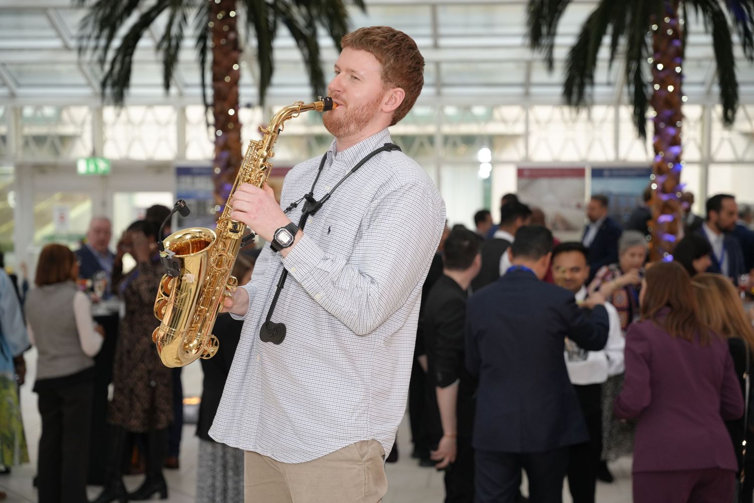 A red-haired man plays alto saxophone at an indoor event with palm trees and guests mingling in the background.