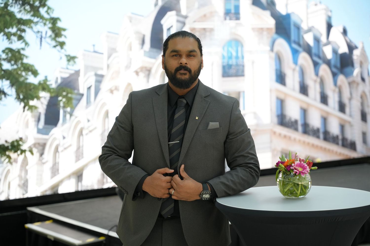 Professional man in gray suit posing outdoors near elegant white building with floral centerpiece.