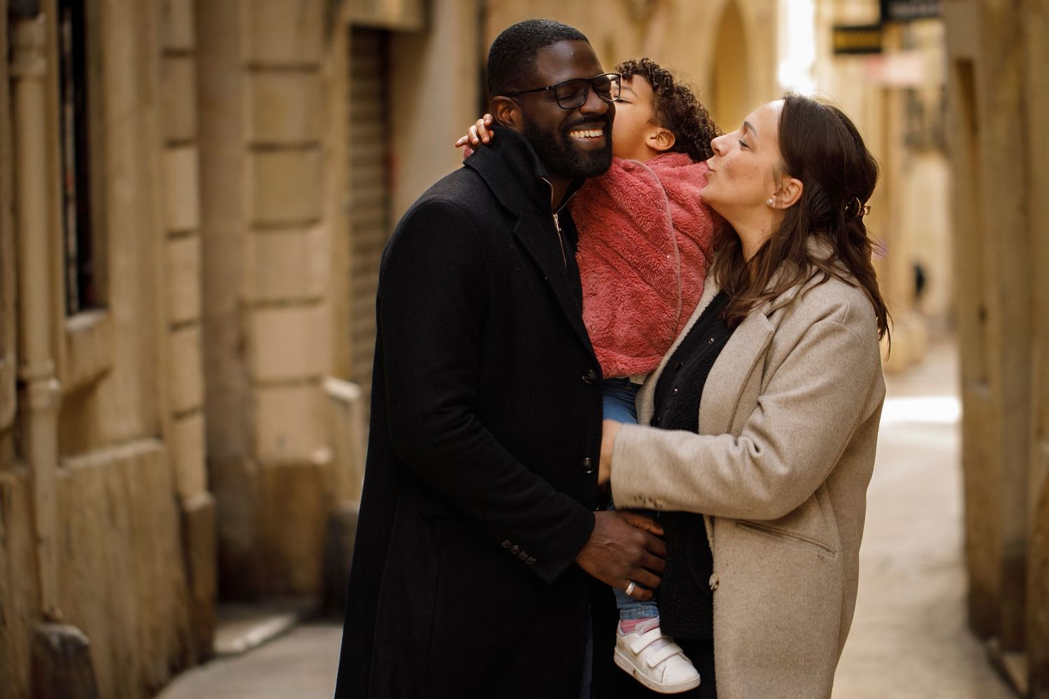 Photo de famille avec un papa, une maman qui rient et la petite fille qui fait un bisou à son papa dans une rue de Montpellier