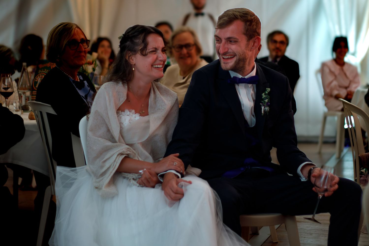 Deux mariés rient aux éclats, assis face à un discours.