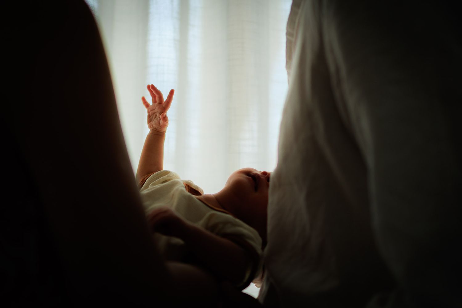 Un bébé dans les bras de ses parents étire son bras vers la lumière d'une fenêtre lors d'une seance naissance à Castelnau le lez.