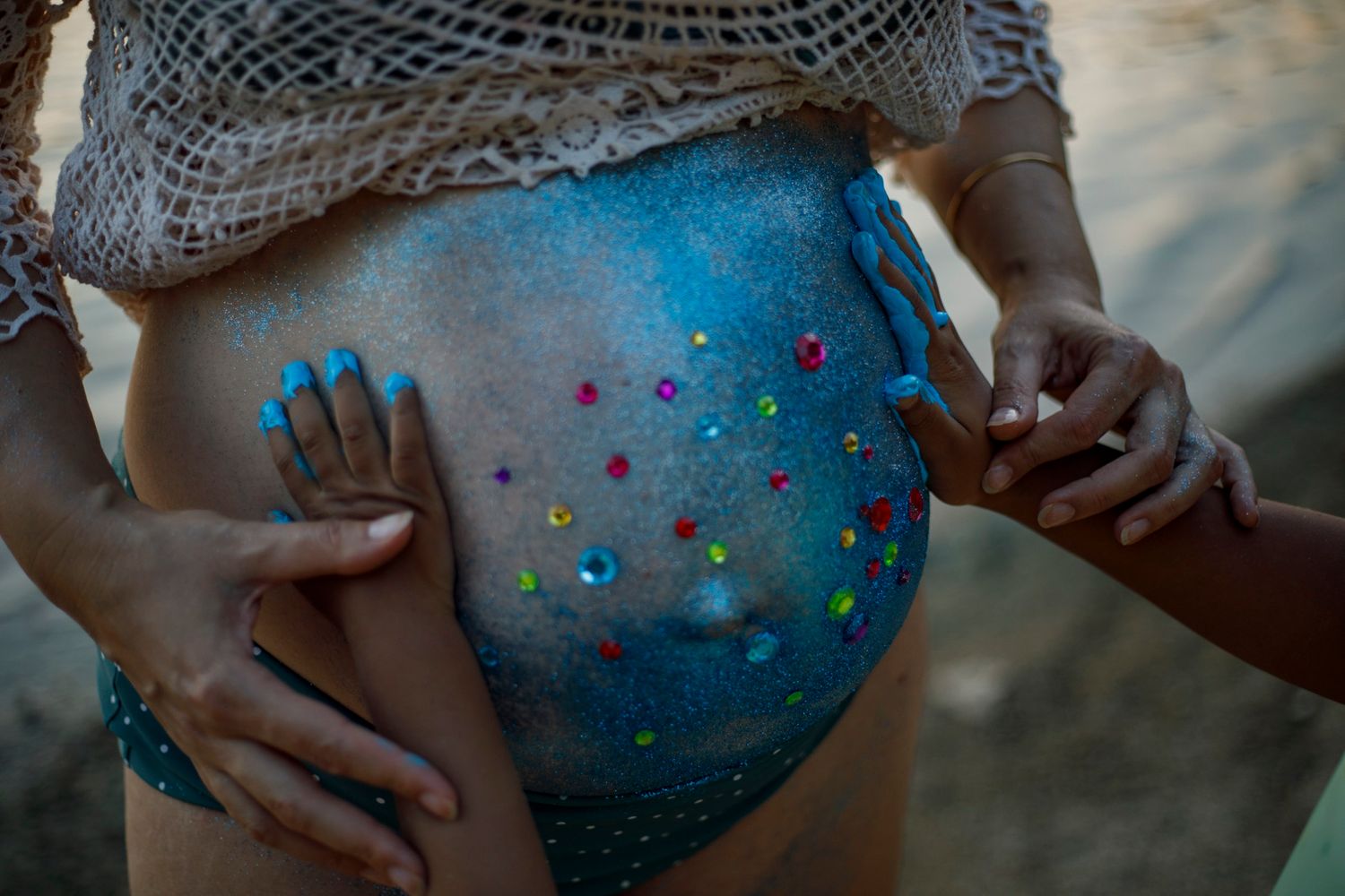 Gros plan sur un ventre de grossesse recouvert de peintures colorées et de paillettes, deux mains d'enfant posées de chaque coté du ventre.