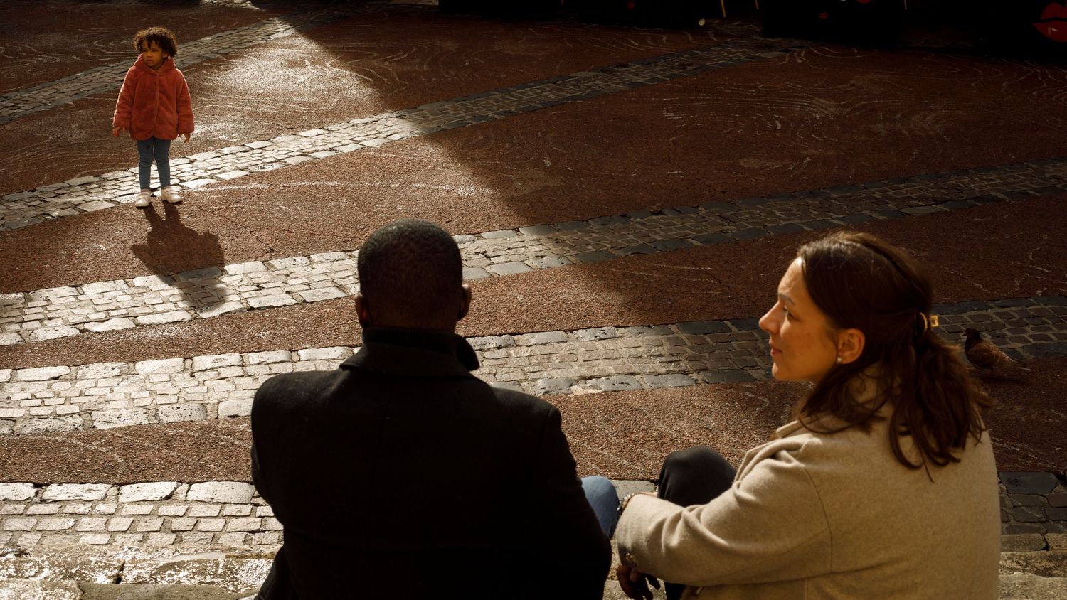 Photo d'un papa et d'une maman qui regardent leur fille jouer dans la lumière qui se fraie un chemin entre les immeubles de l'écusson