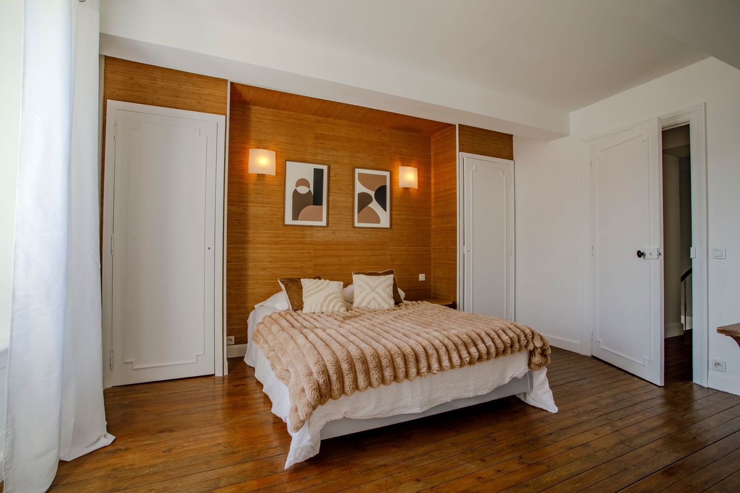 Une chambre d'un gîte de charme avec un grand lit double recouvert d'un plaid taupe, une tête de lit en bois et des murs blancs.