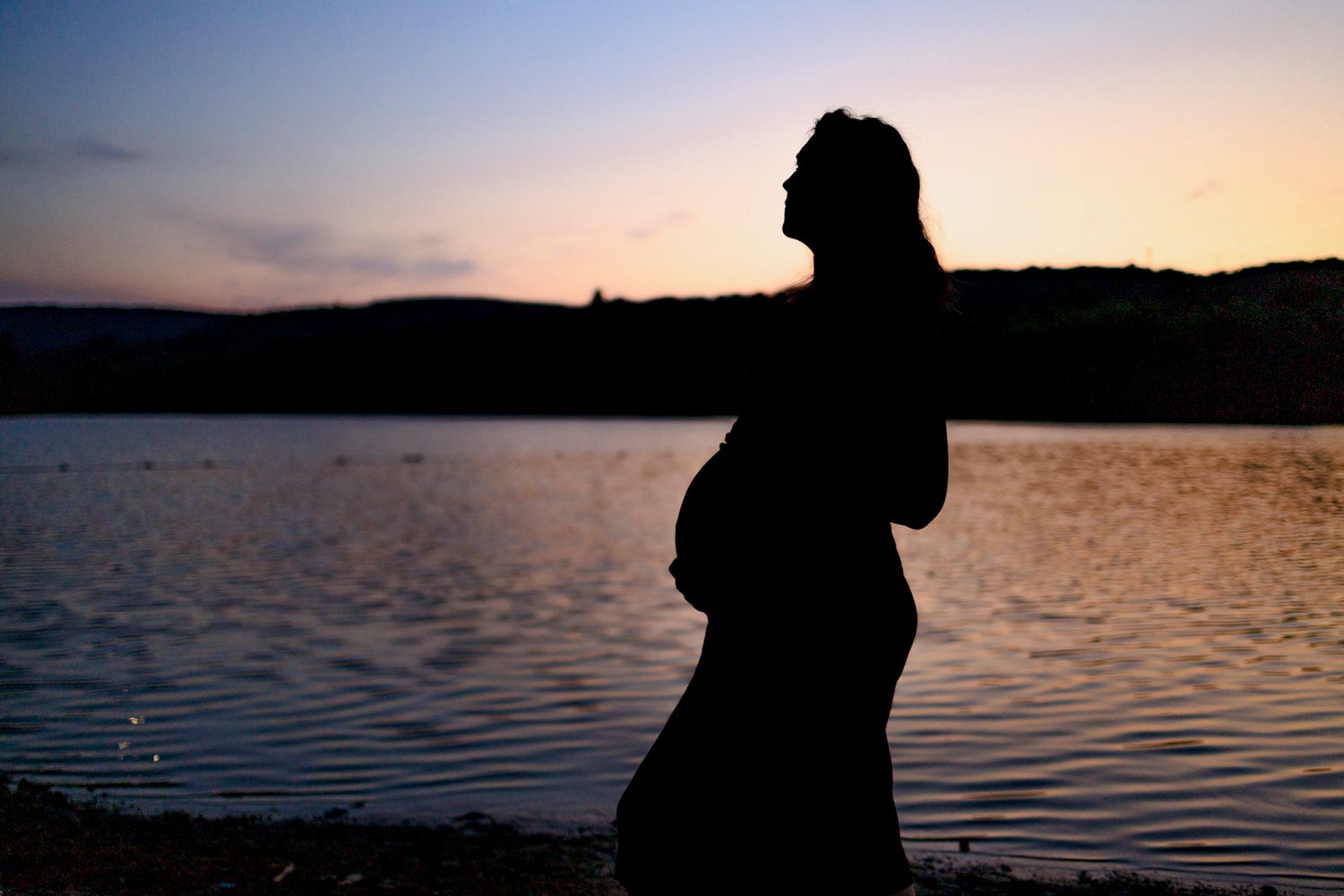 Silhouette d'une femme enceinte devant un lac au soleil couchant.