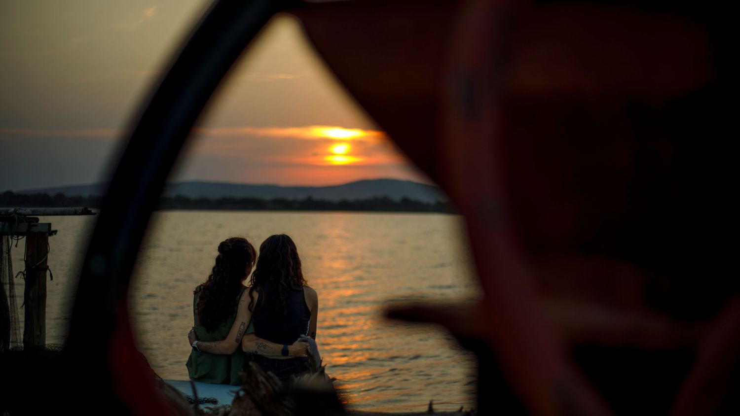 Photo d'un couple de femme de dos, assises bras dessus bras dessous face au coucher de soleil au bord de l'Etang des Mourres