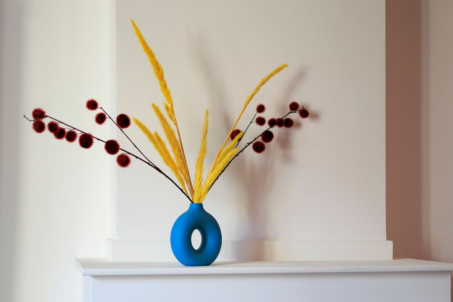 Détail décoratif élégant avec un arrangement de fleurs séchées jaunes et rouges dans un vase bleu moderne, posé sur une cheminée.