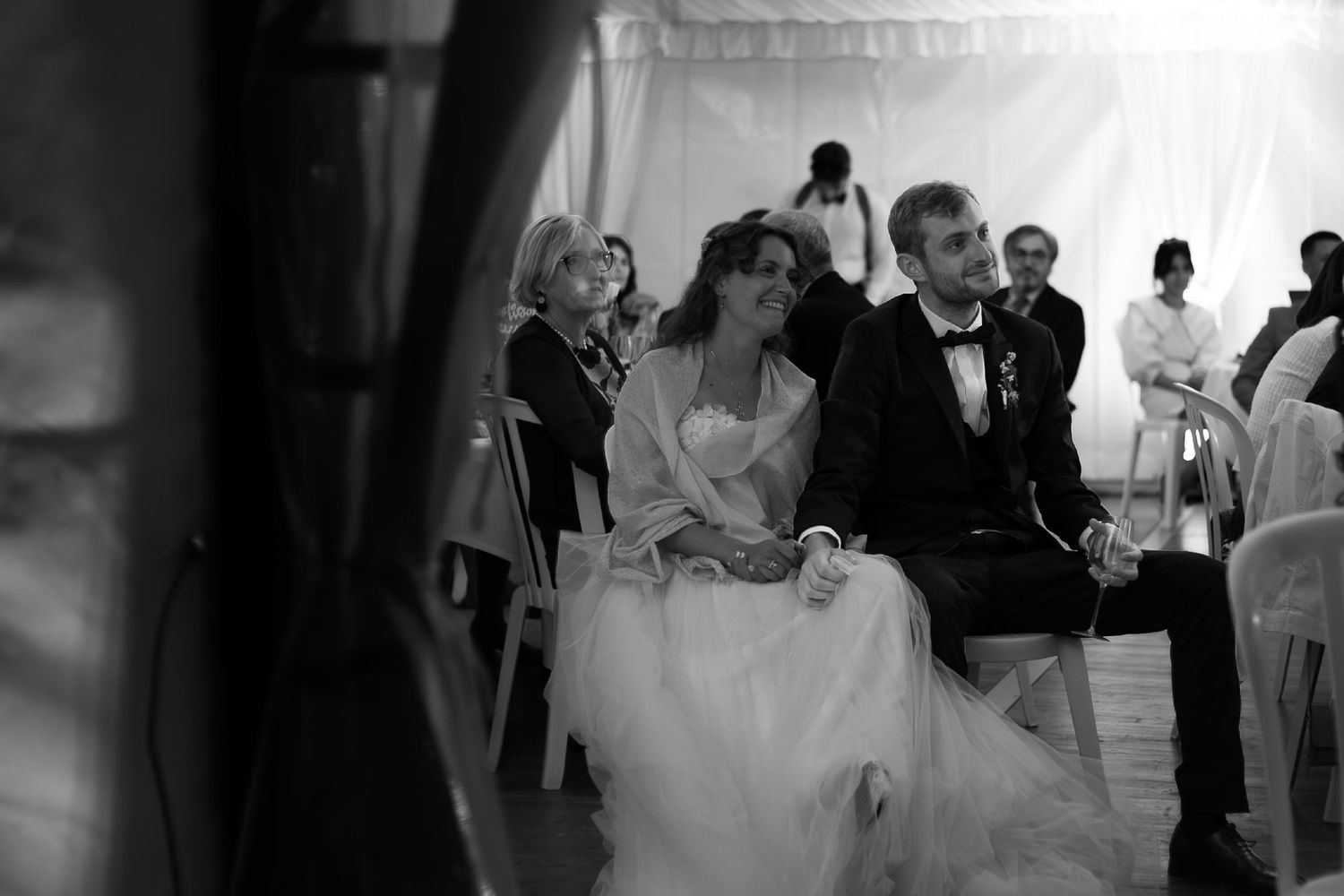 Photo noir et blanc montrant des mariés assis écoutent les discours lors d'un reportage mariage.