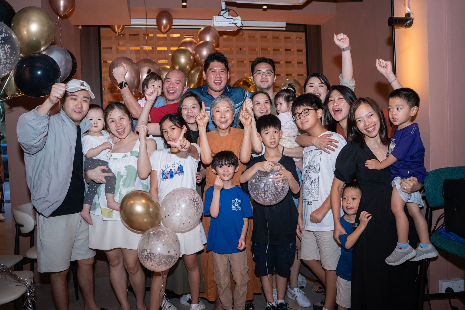 Large Asian family group celebrating a party with balloons, raised arms, and big smiles in a festive indoor setting.