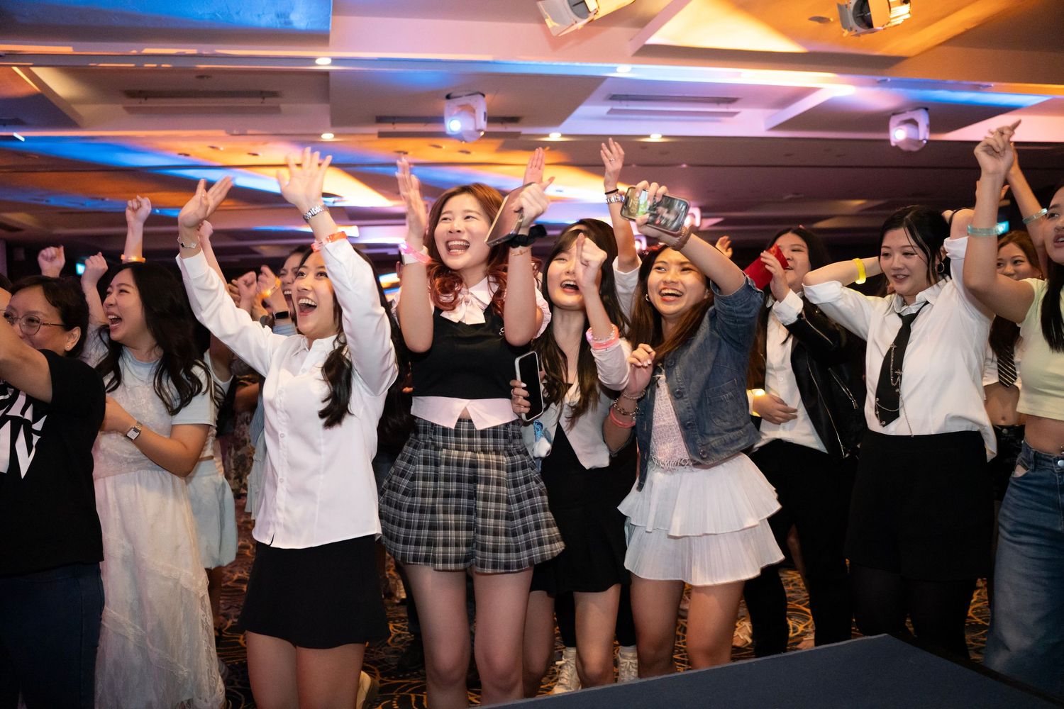 A crowd of party-goers dancing and celebrating with raised arms at a festive indoor event with colorful lighting.