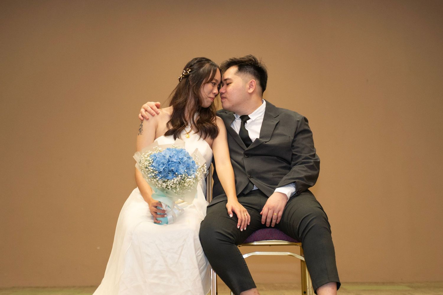 Studio portrait of couple in wedding attire with blue bouquet against brown backdrop.