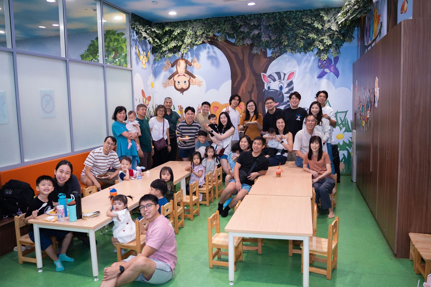 Group of adults and children posing together in a colorful kids classroom with a tree mural wall.