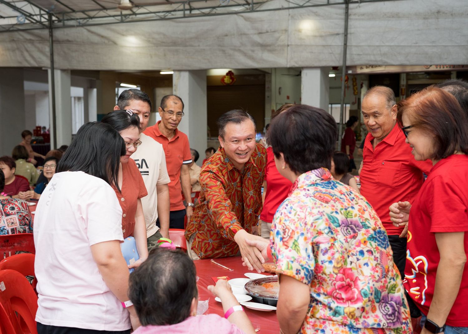Man in red shirt greeting elderly guests at a community gathering event in a decorated hall.