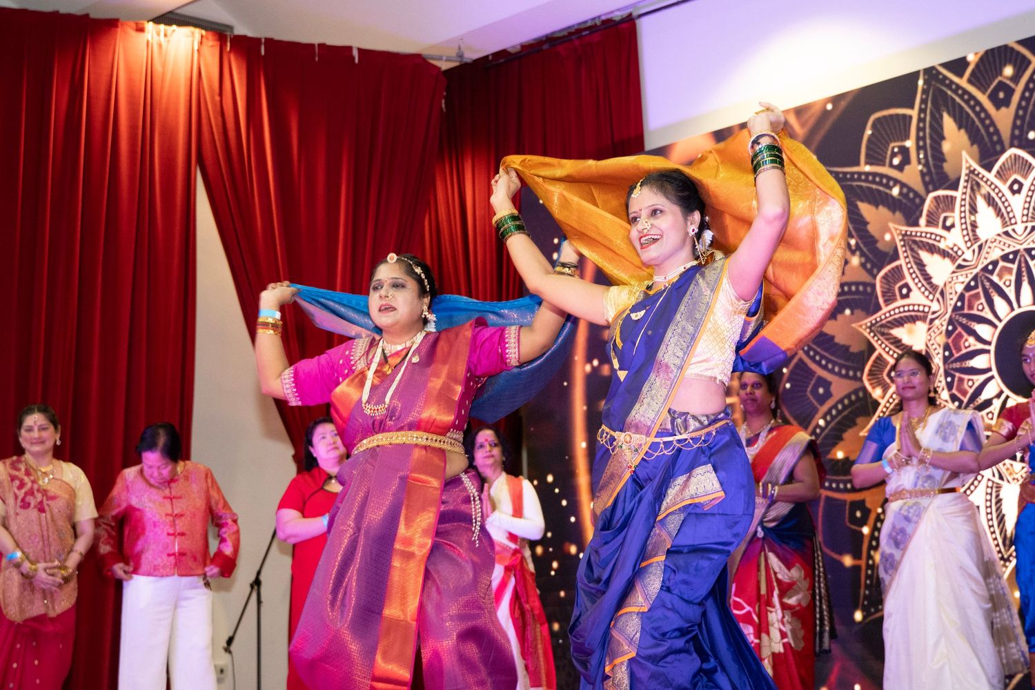 Colorful cultural dance performance featuring traditional costumes and choreography on a stage with red curtains.