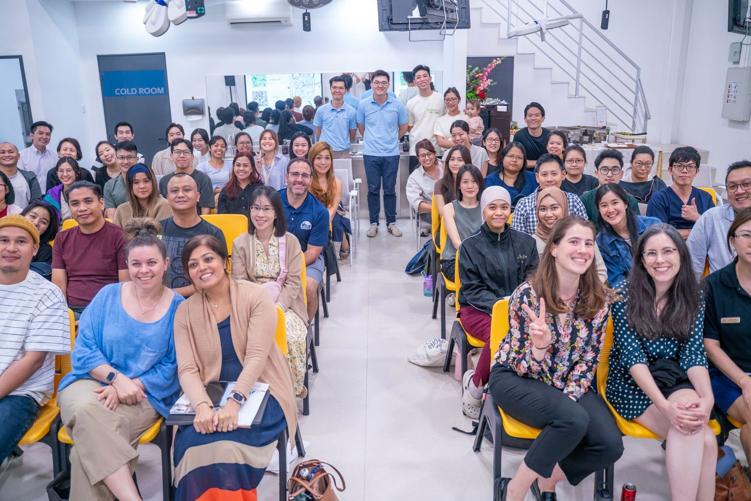 Large group of diverse people posing for a photo in a modern seminar room with yellow chairs.