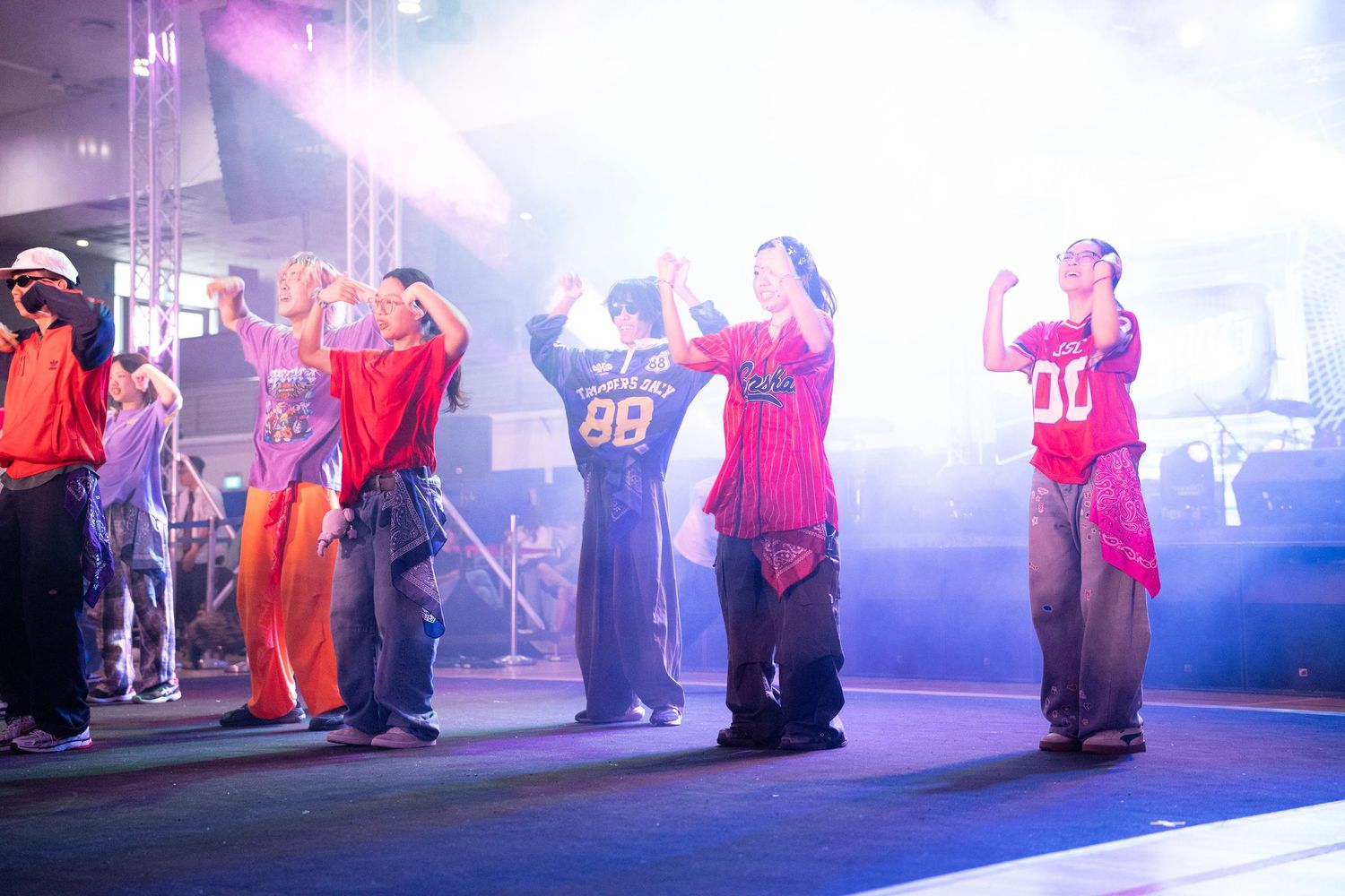 Performers in colorful streetwear dance on a foggy stage under bright lights at a live event.
