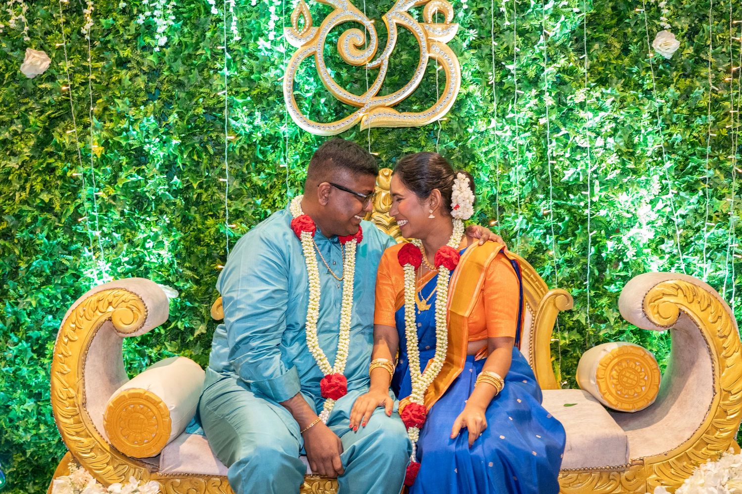 Couple sharing intimate moment on decorative throne with green floral backdrop.