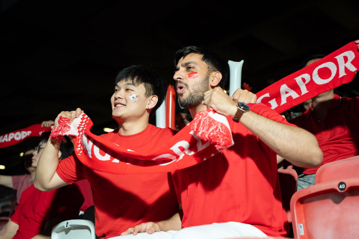 Two Singapore football fans in red jerseys cheering with a Singapore scarf at a stadium.