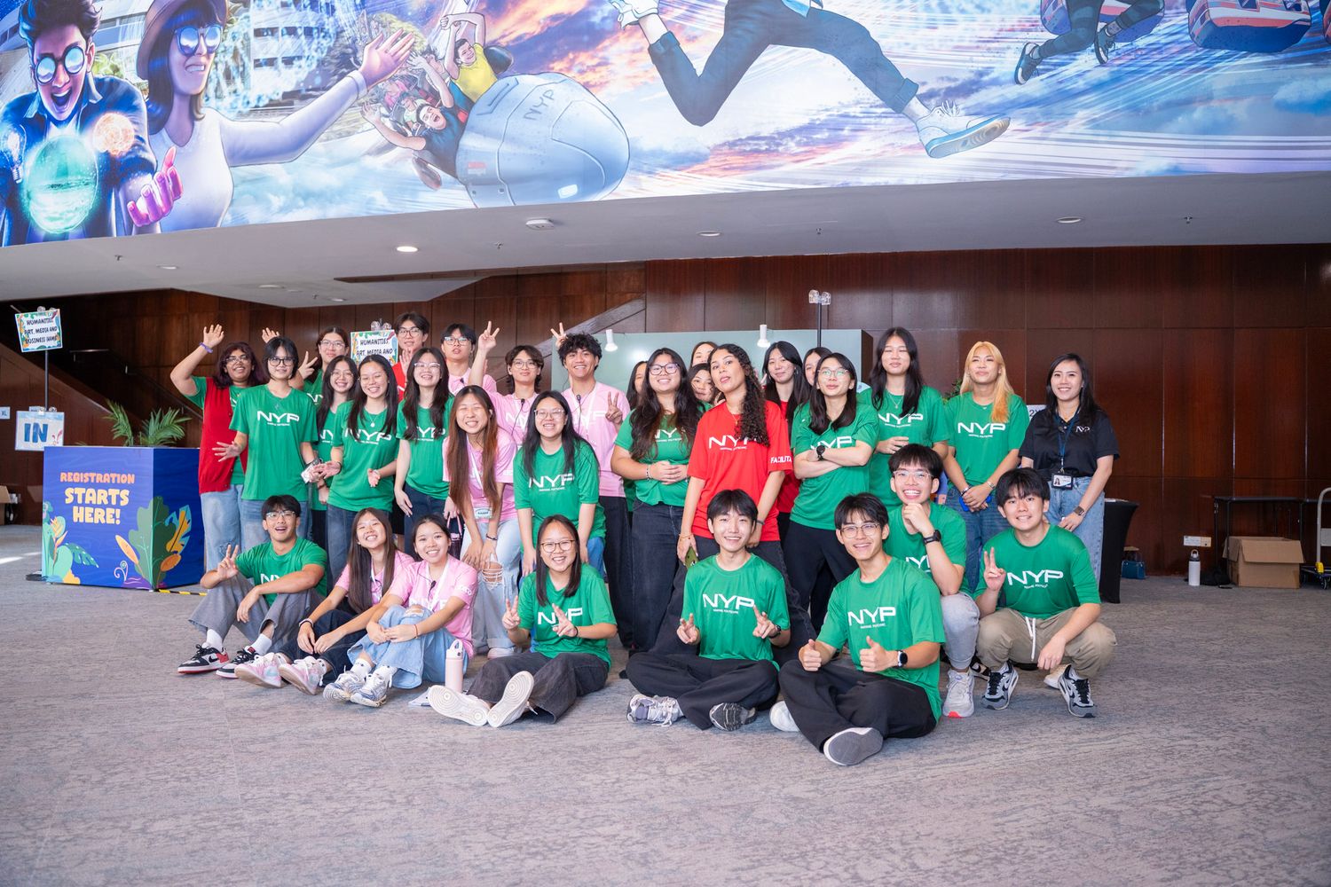 Large group of students in green shirts posing together in a modern venue lobby with colorful movie posters.