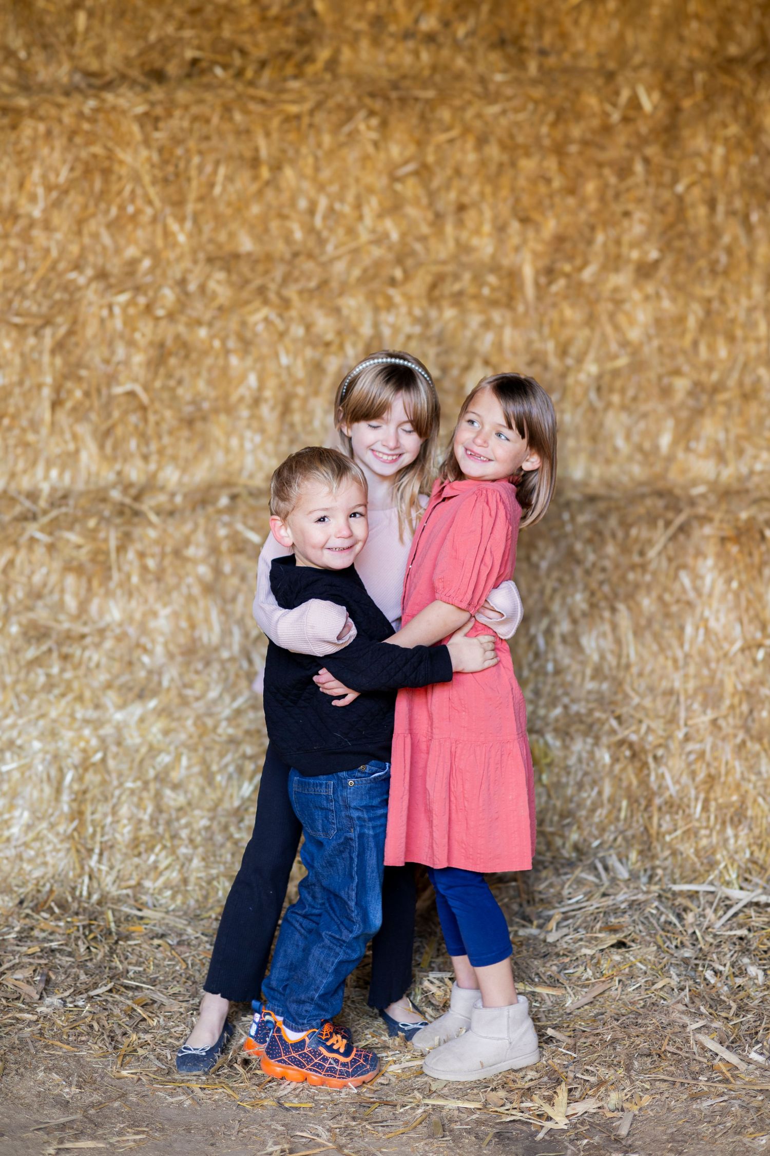 Three children pose together against a hay bale background in casual clothing.
