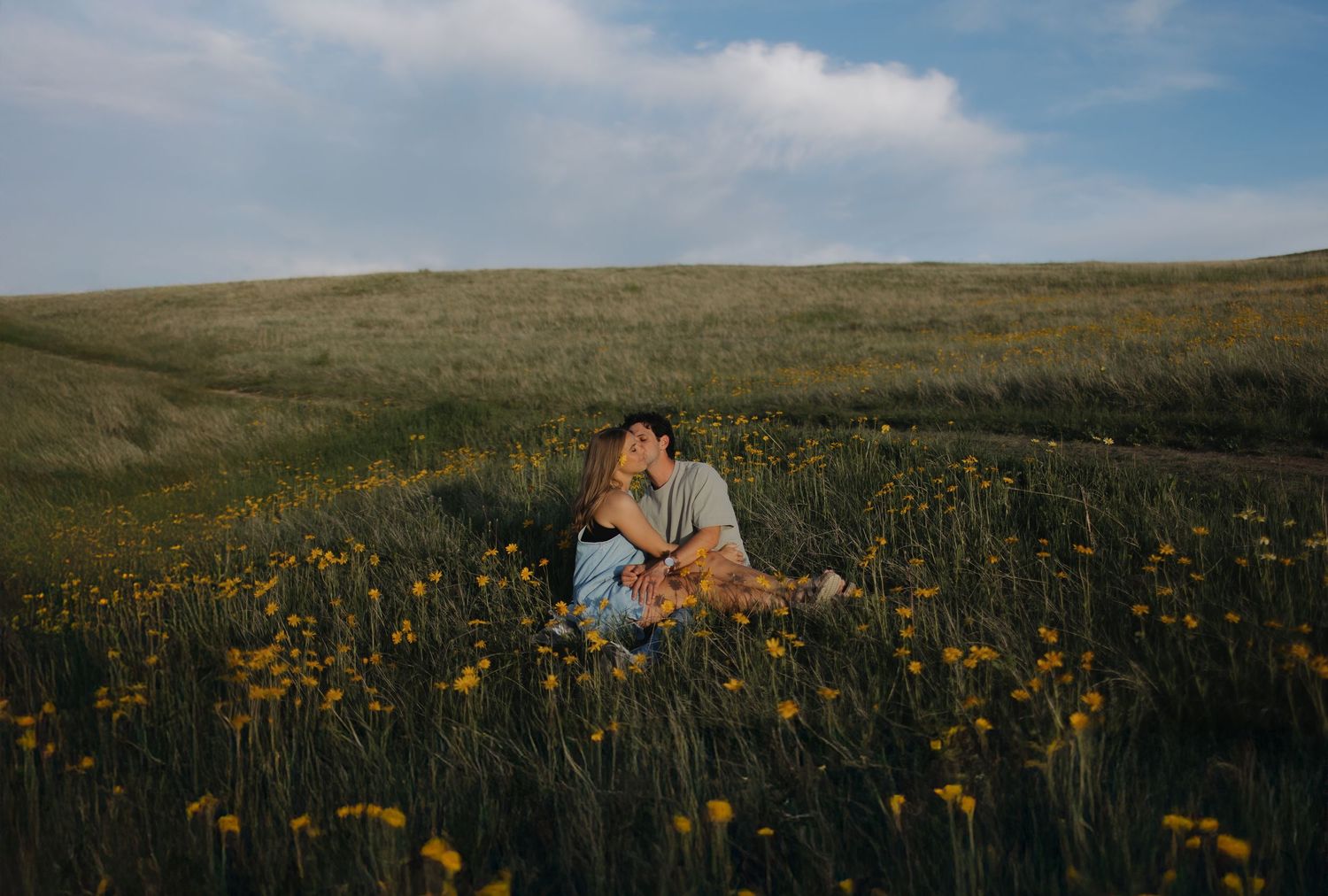 Couple posing in field of yellow wildflowers with mountains in background.