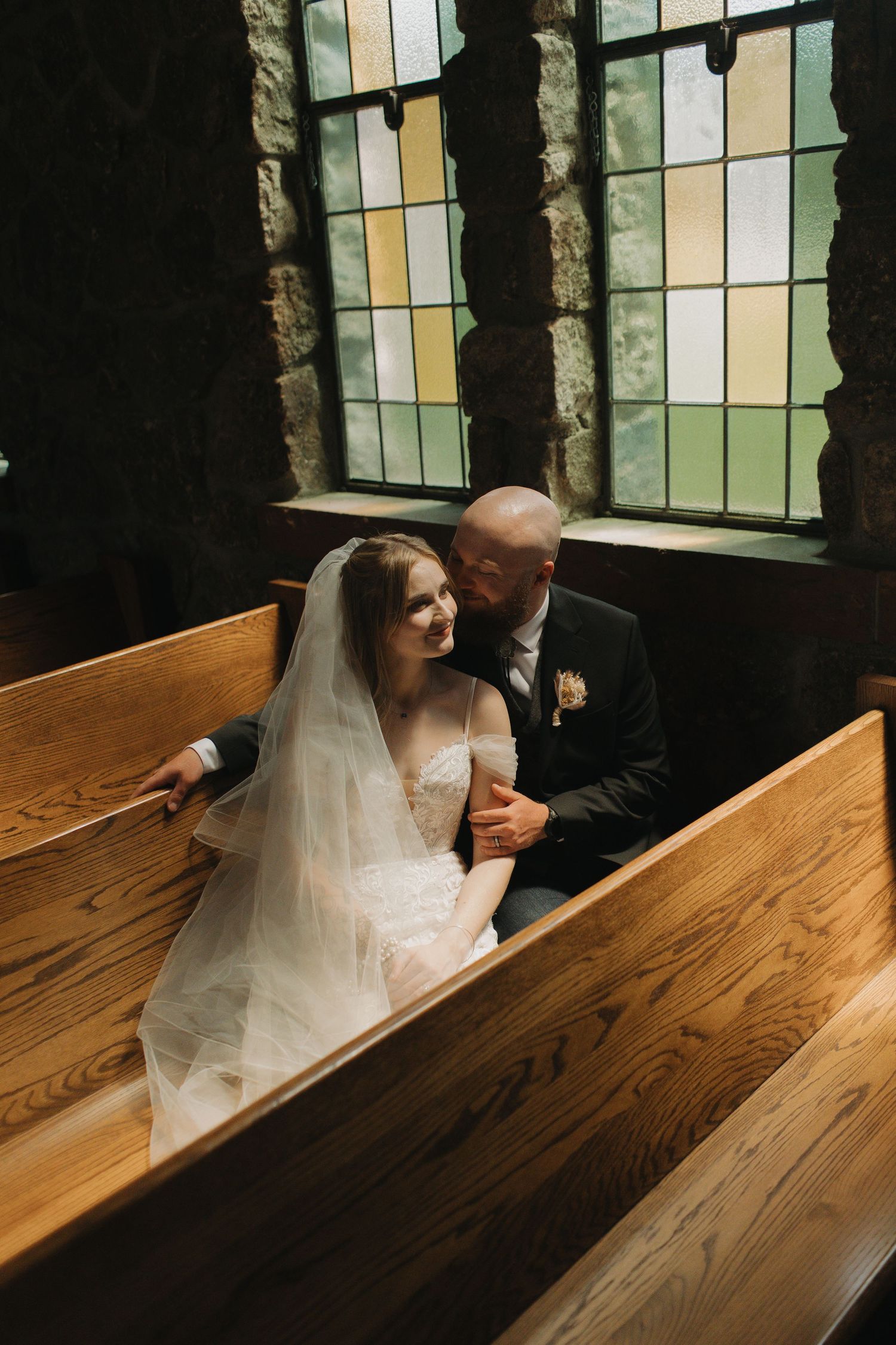 Intimate wedding moment captured in wooden church pews with sunlight streaming through stained glass windows.