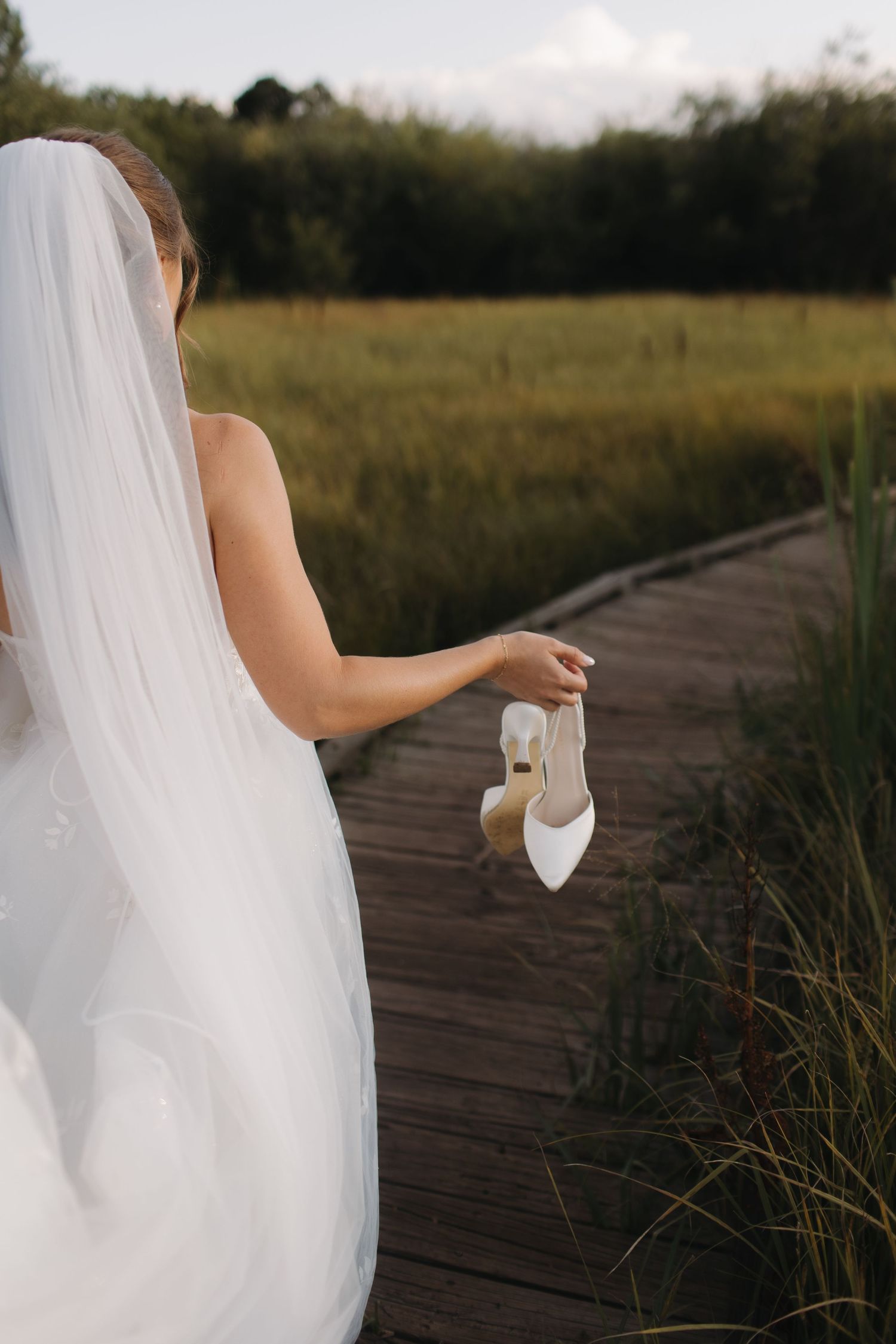 Wedding dress and veil details captured on a wooden boardwalk surrounded by tall grass.