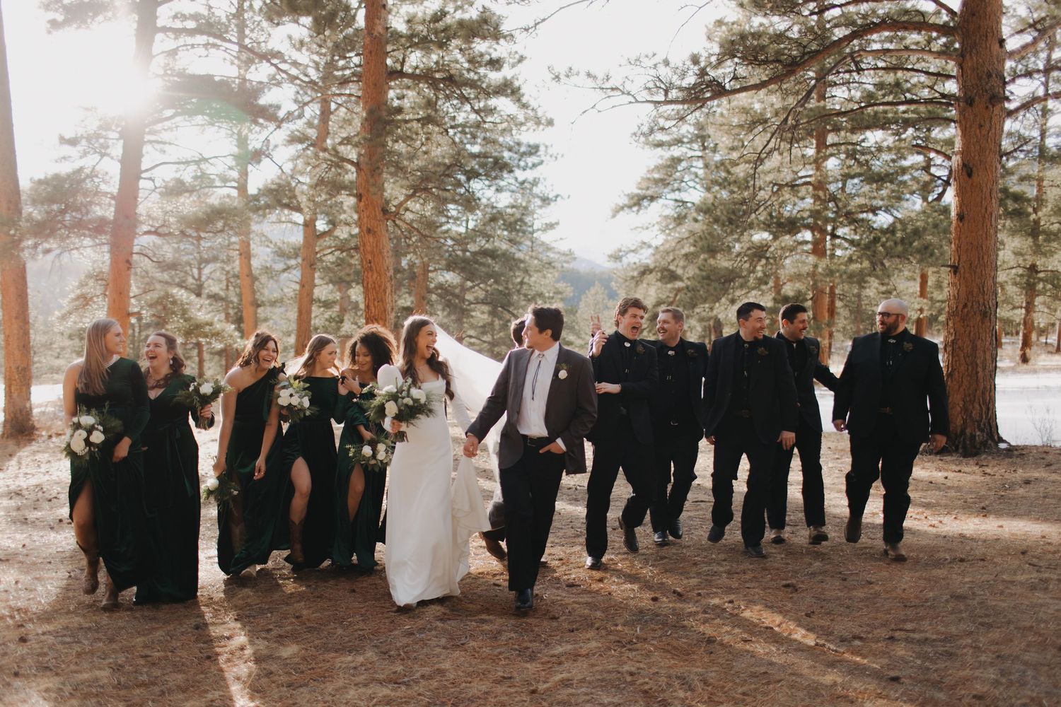 A joyful wedding party in black attire runs through a pine forest on a sunny day.