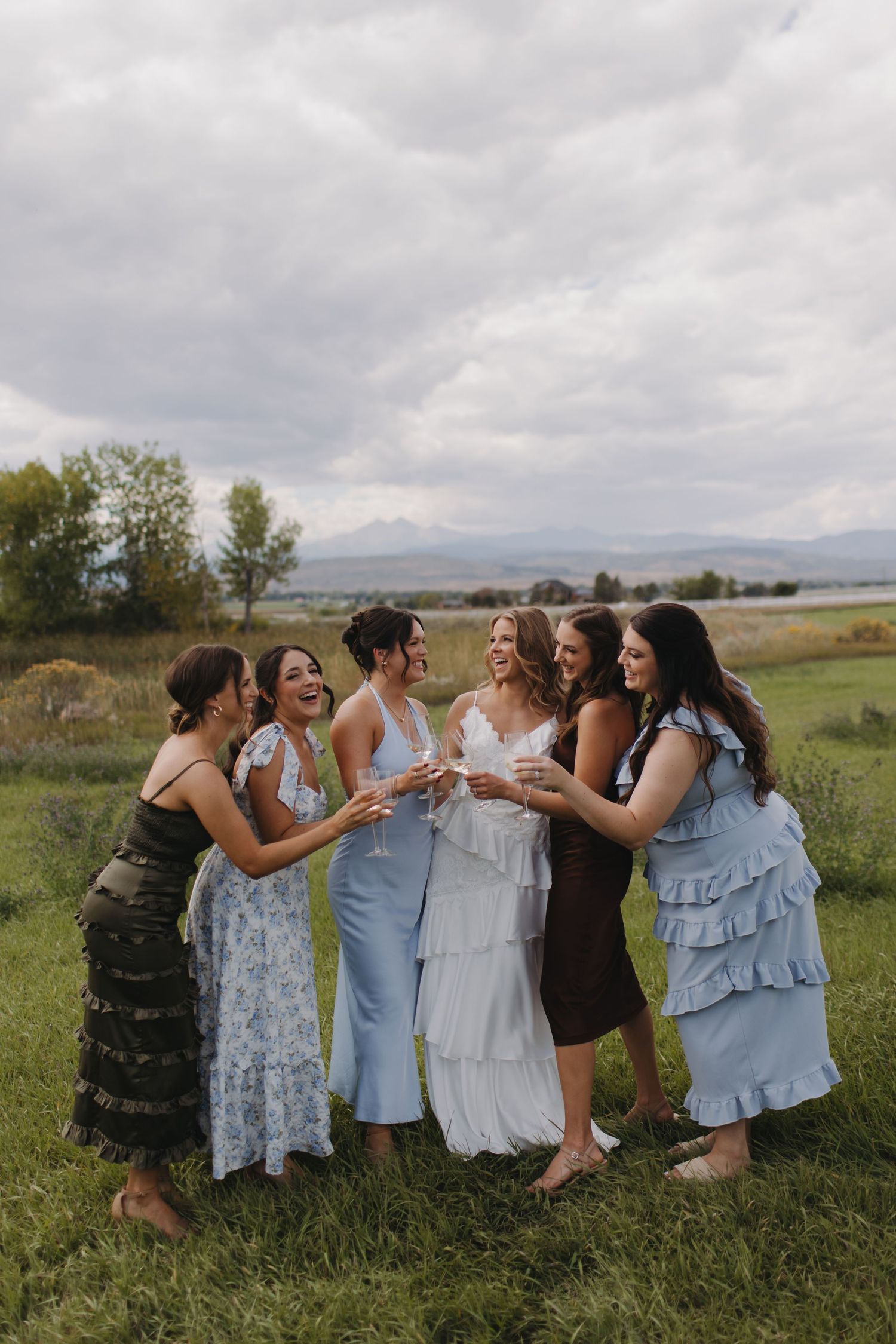 A group of wedding guests in flowing dresses gather on grassy hills with mountains in the background.