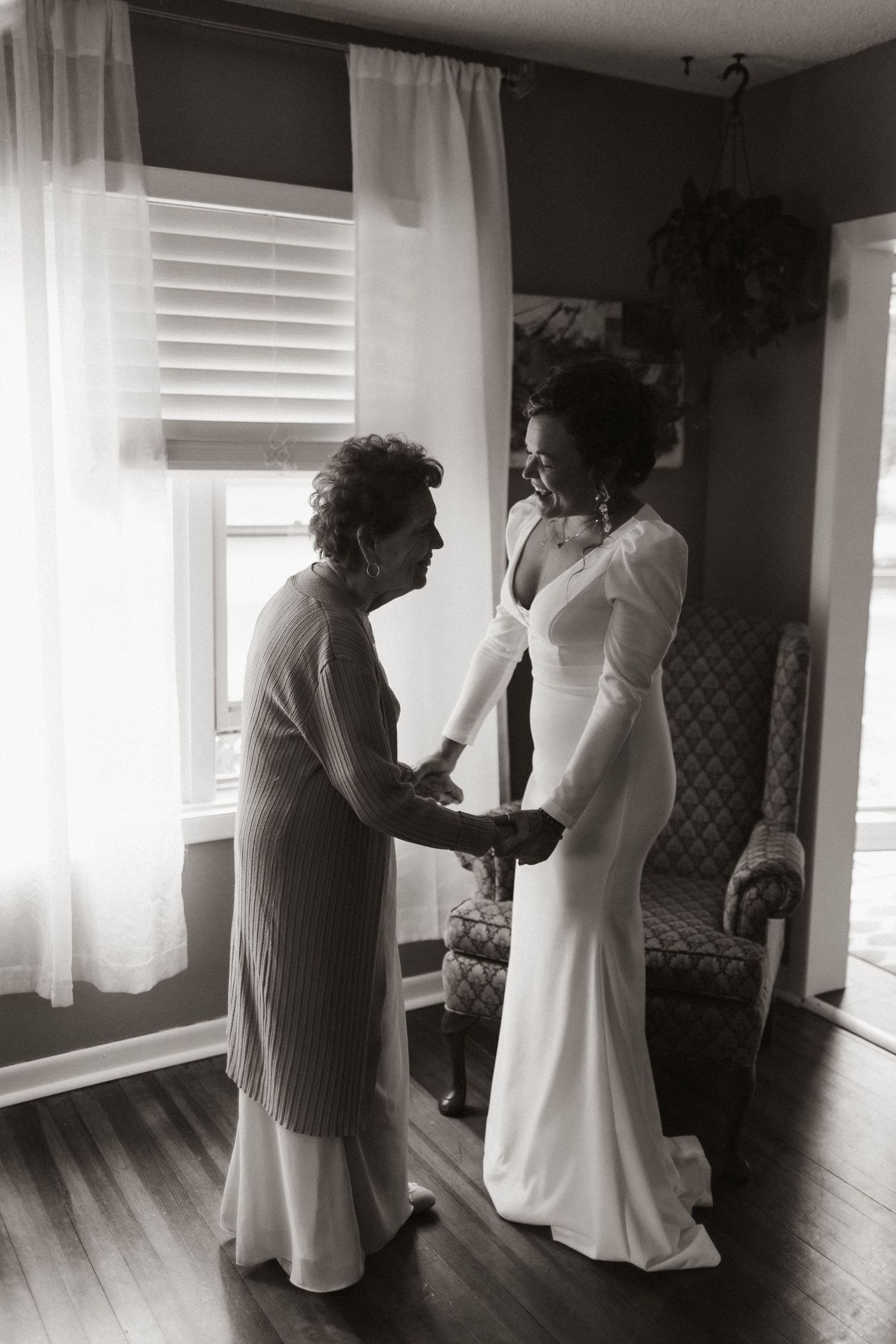 A touching black and white moment showing wedding dress preparations in a sunlit room with sheer curtains.