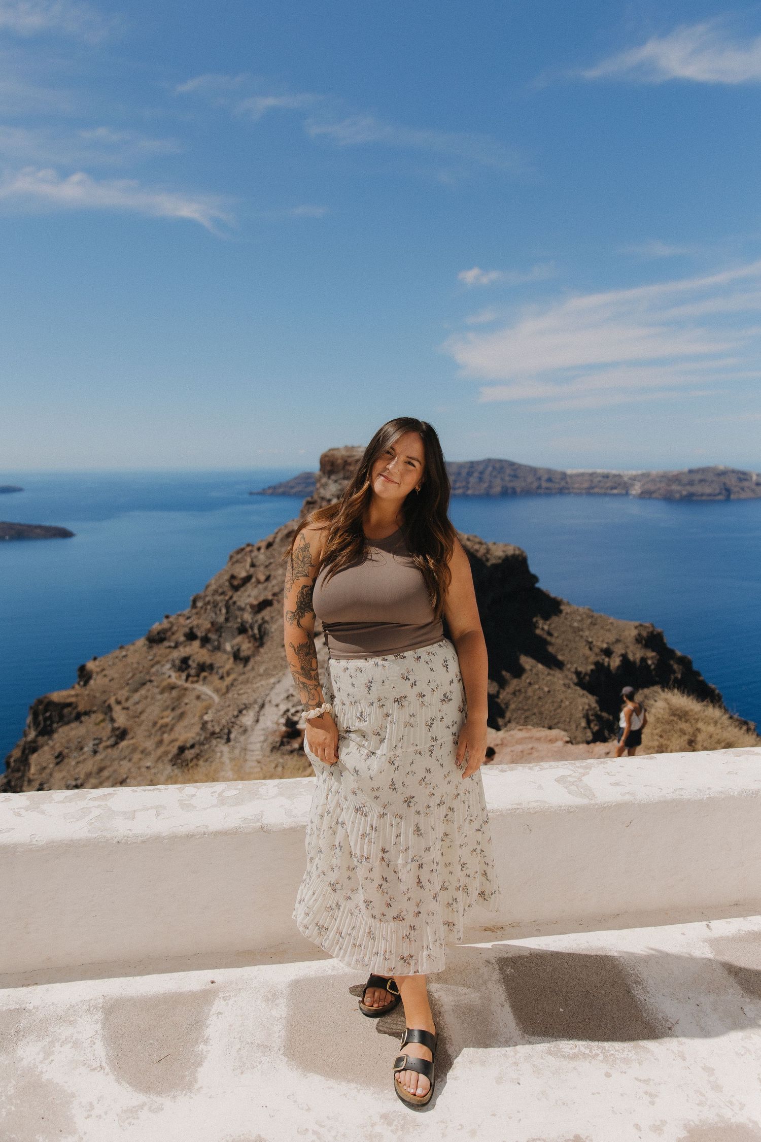 Woman in floral dress leaning on white wall overlooking Mediterranean Sea and islands.