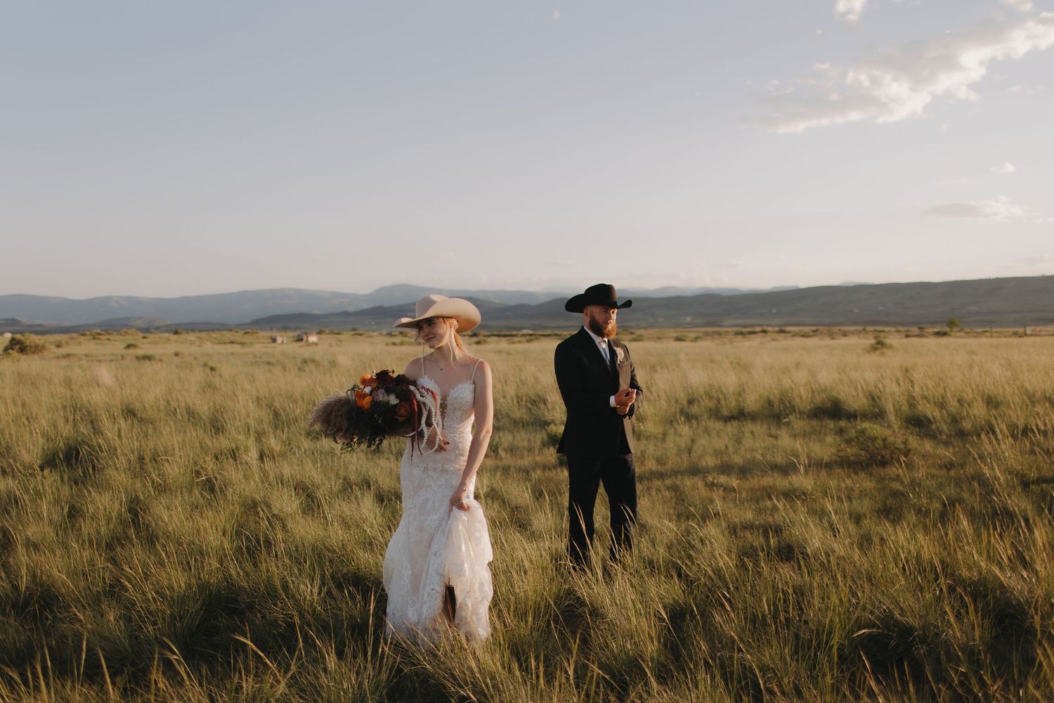 Couple walking through tall grass in an open field at sunset with mountains in the distance.
