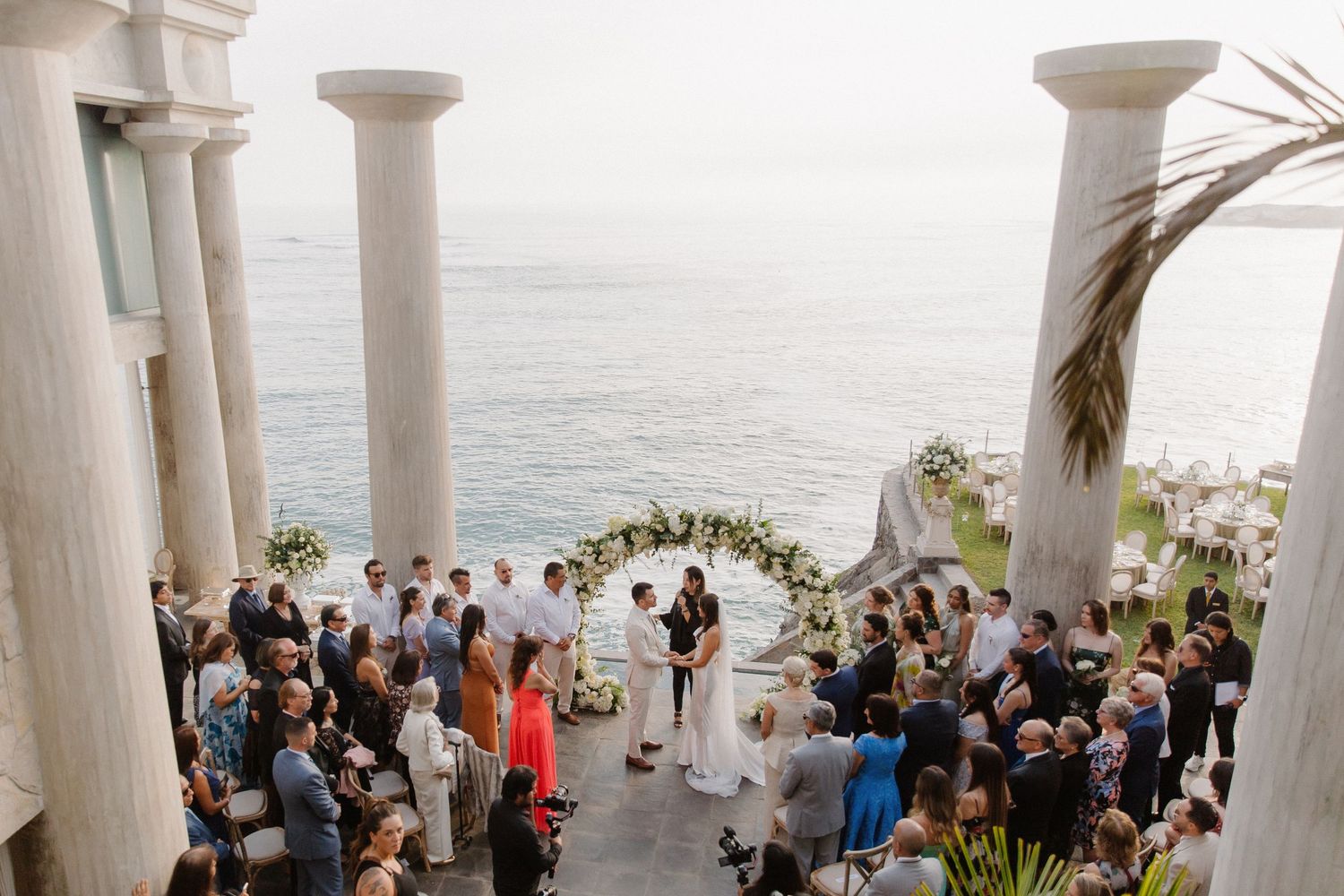 Elegant outdoor wedding ceremony setup with white columns and ocean view backdrop.