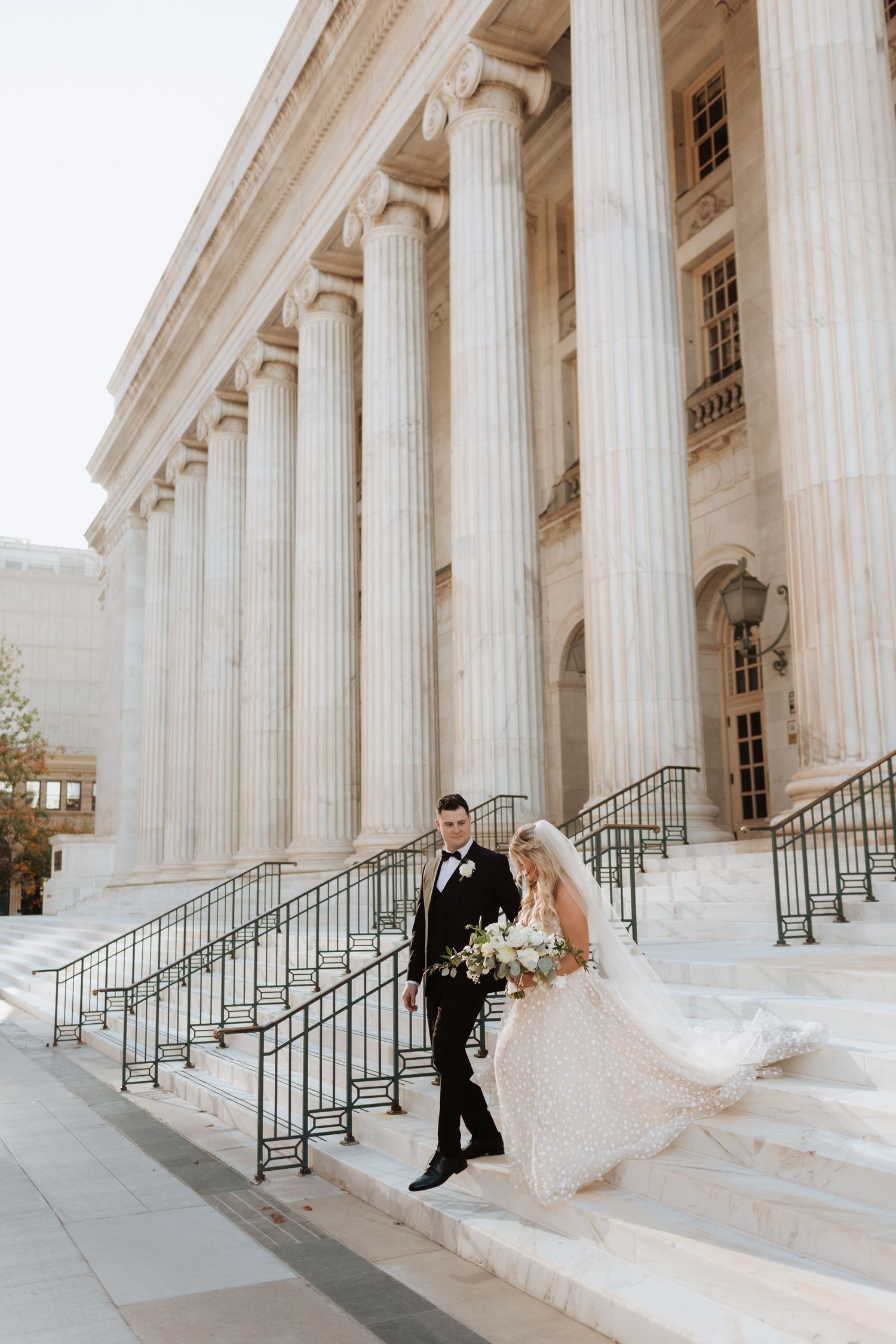 Wedding couple walks along grand neoclassical building with white columns.
