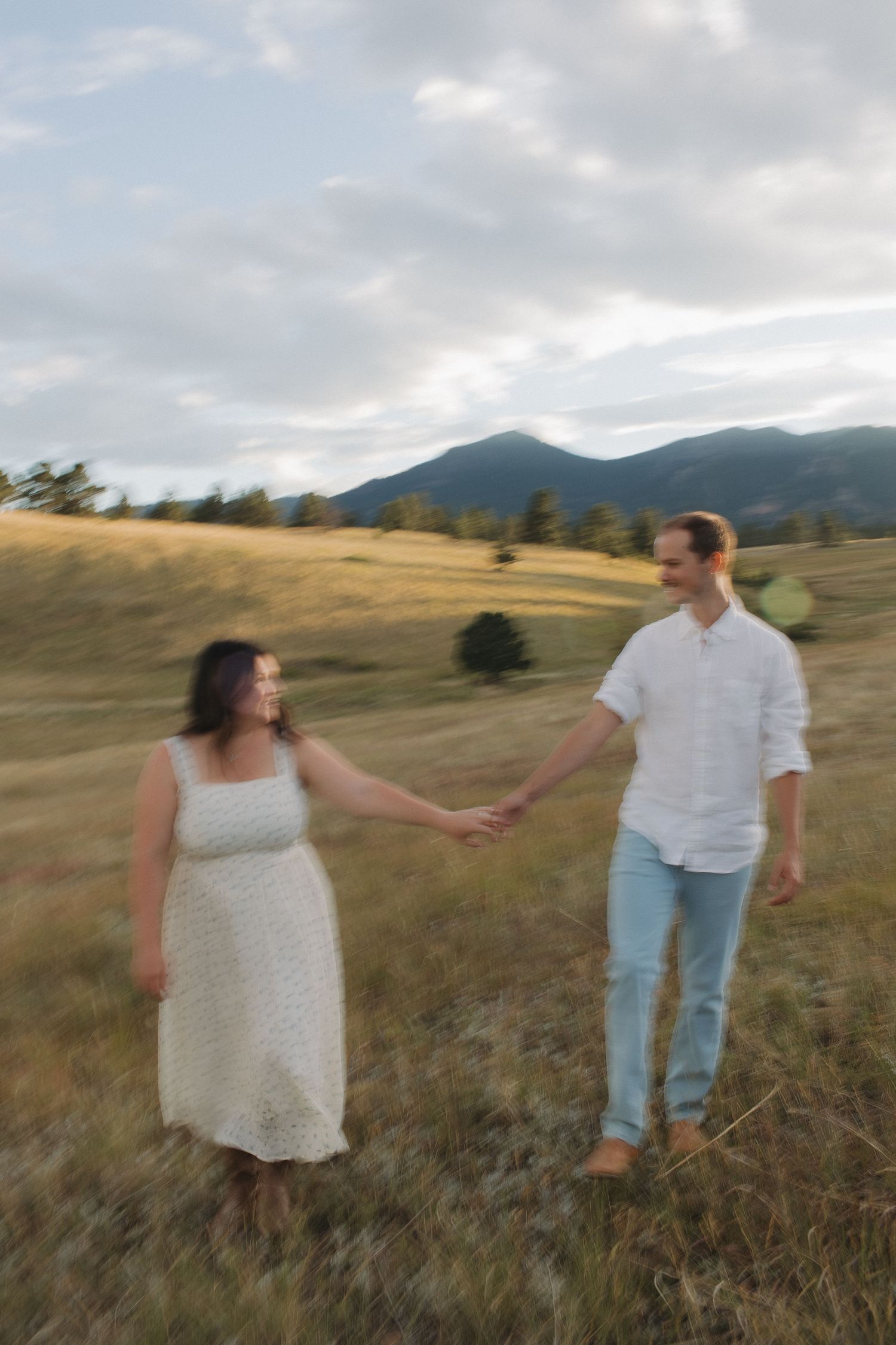 A couple holds hands while walking through a grassy meadow with mountains in the background.
