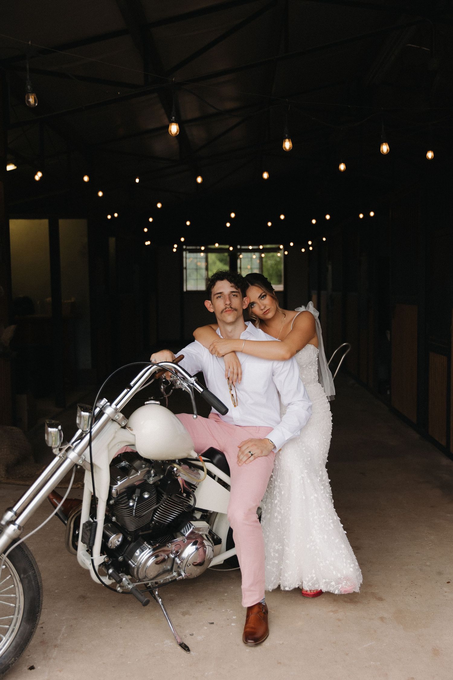 Couple poses with a motorcycle in a rustic venue with string lights overhead during their wedding celebration.