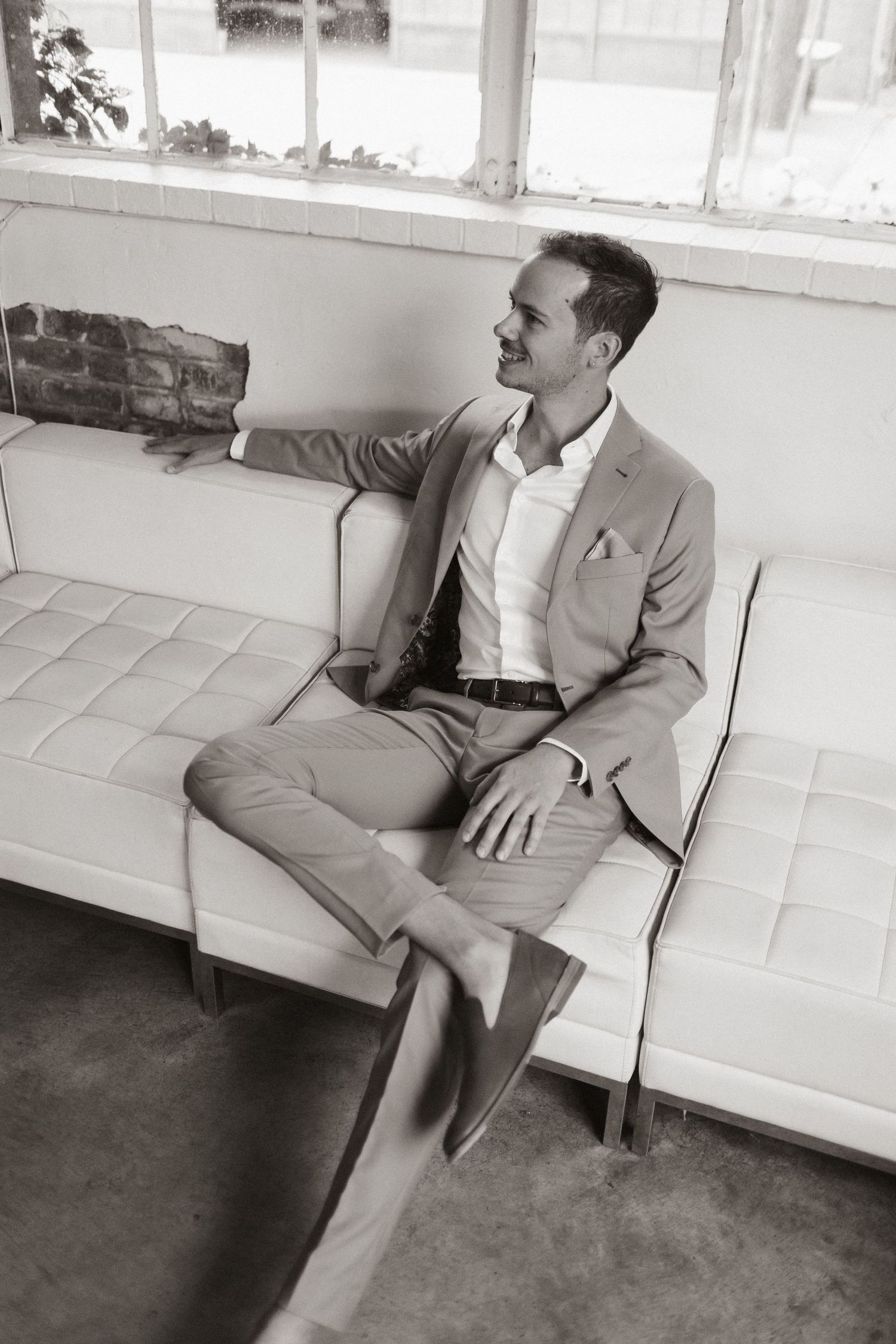 Black and white photo showing a person in a light colored suit sitting on a white couch in a bright room.