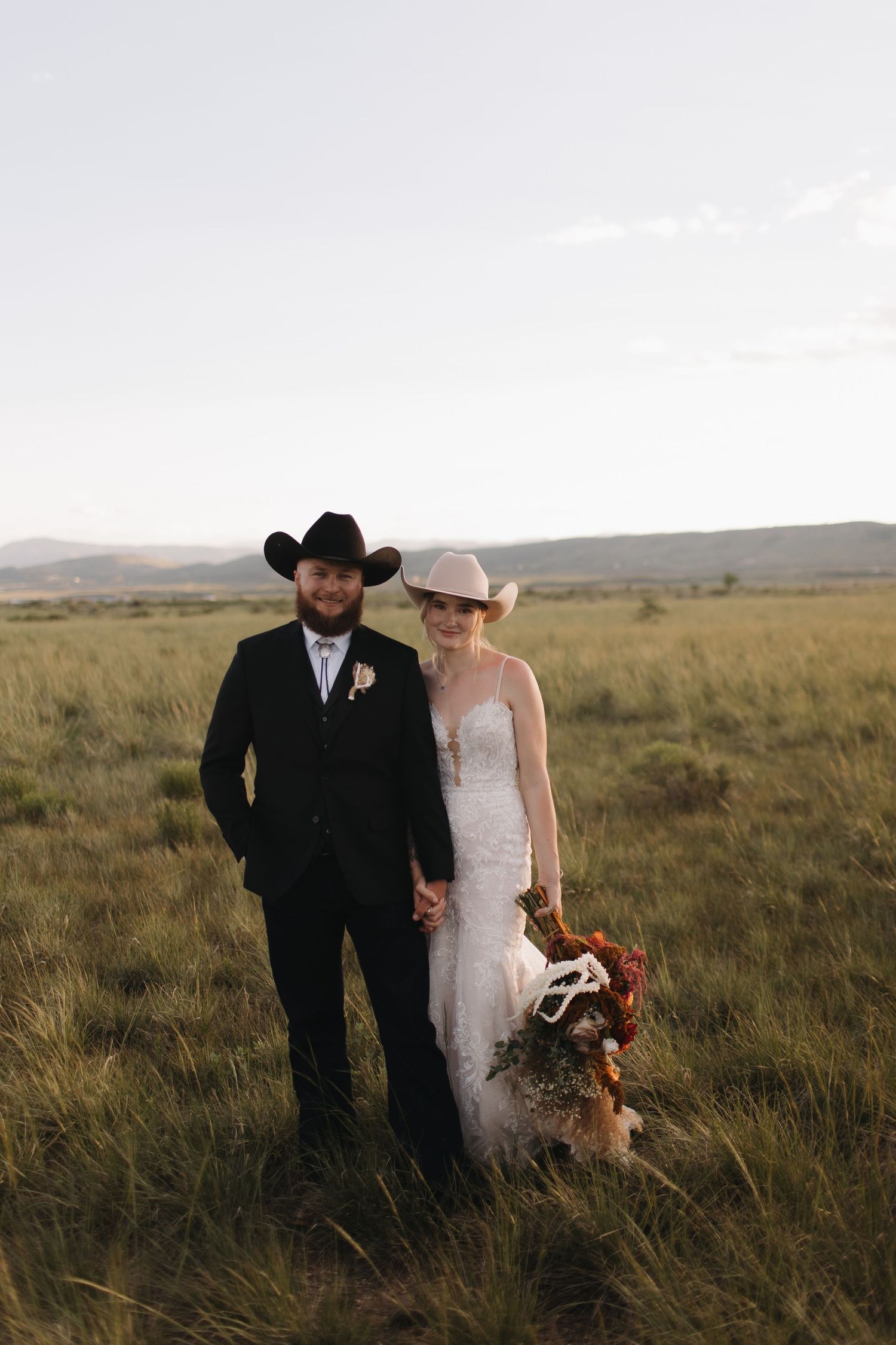 Wedding couple walks through open prairie field at sunset wearing western attire.