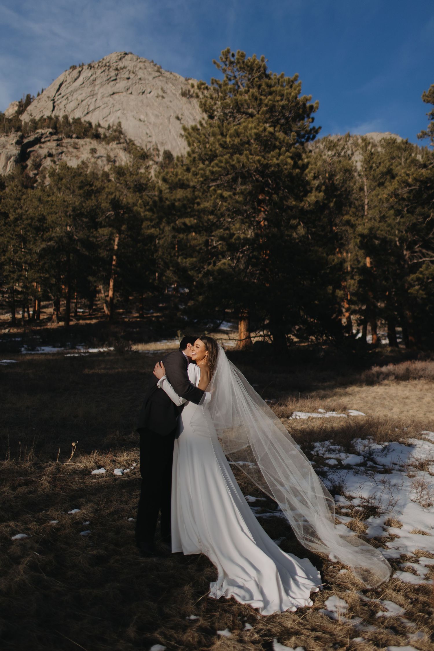 A dramatic wedding portrait against a mountain backdrop with snow patches and evergreen trees.
